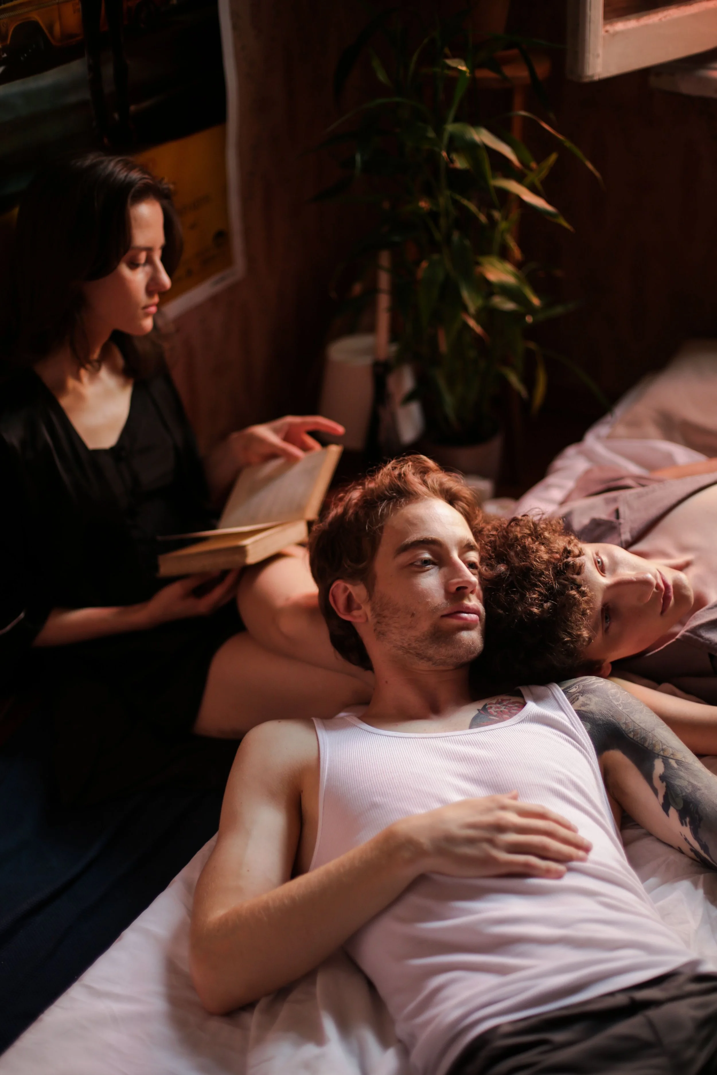 Three young people lying on a bed with two men and one woman. The woman is reading a book and the two men are resting with their eyes closed. The setting appears to be a cozy, dimly lit room with a large potted plant and various objects nearby.