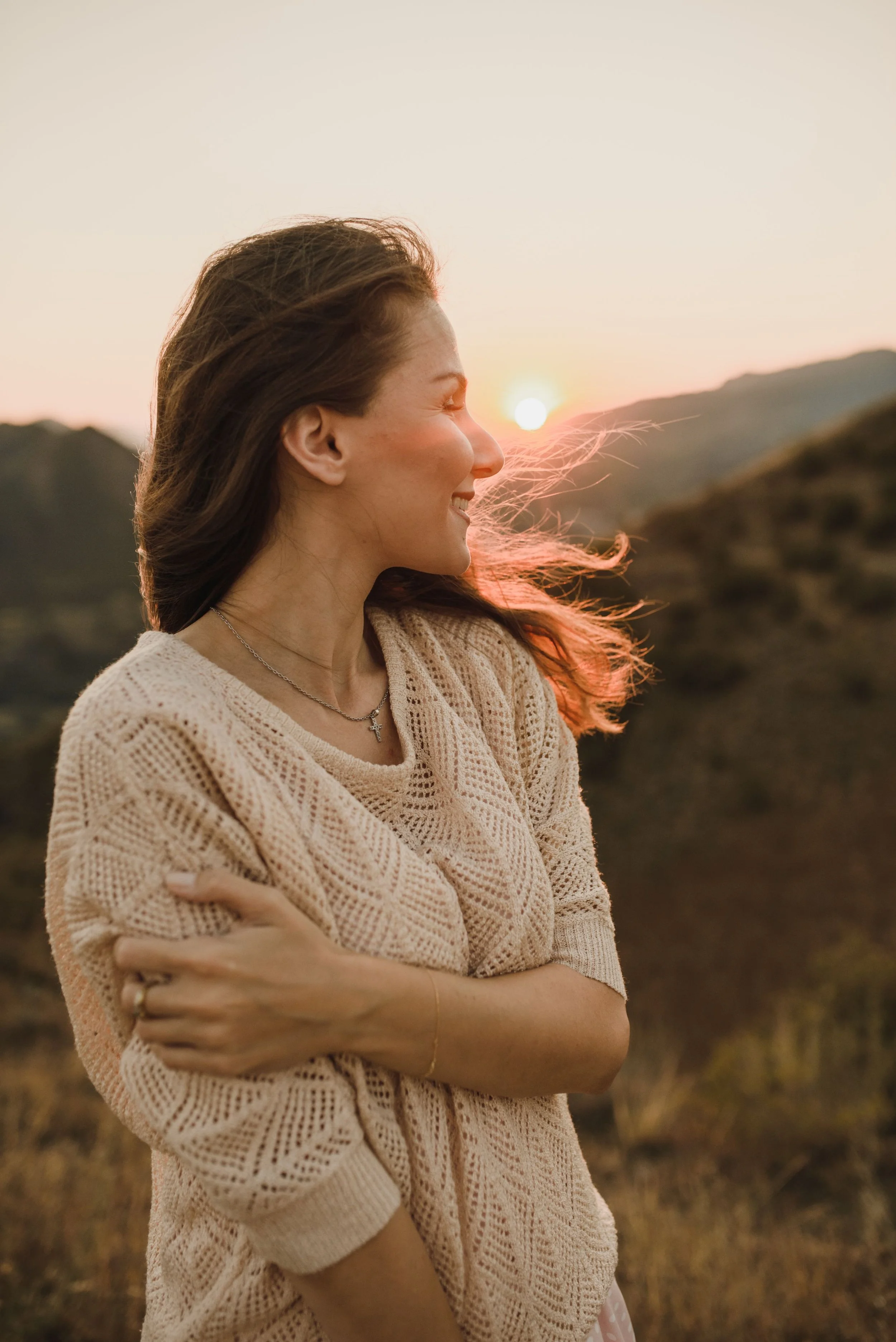 A woman with brown hair smiling with her eyes closed, wearing a beige knitted sweater and a necklace with a cross pendant, standing outdoors during sunset or sunrise in a mountainous area.