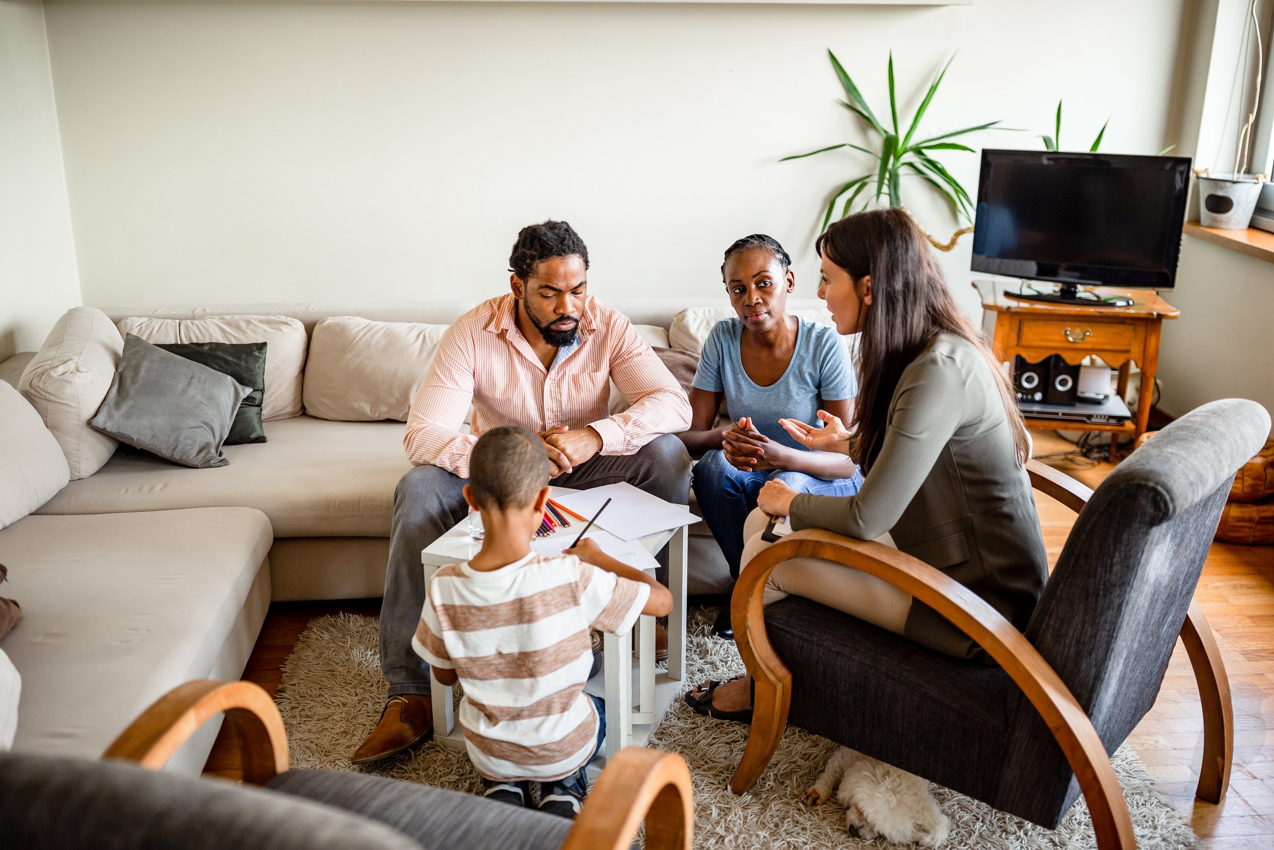 Family of four with two children sitting around a small white table, engaging in conversation in a cozy living room with beige and black sofas, a television on a wooden stand, a tall green plant, and a dog lying on the rug.