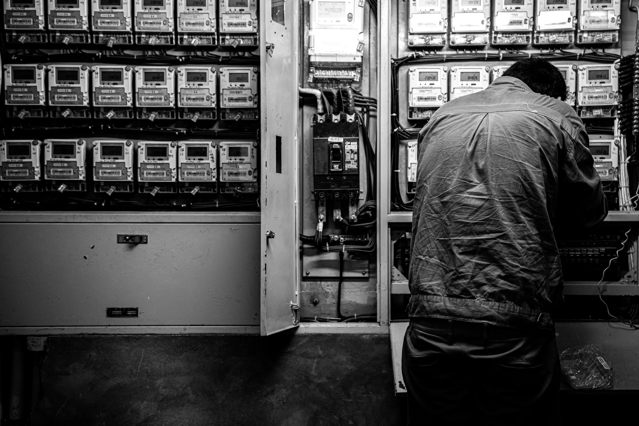 A person in work attire working inside an open electrical control panel or circuit breaker box, with multiple meters and wiring visible.