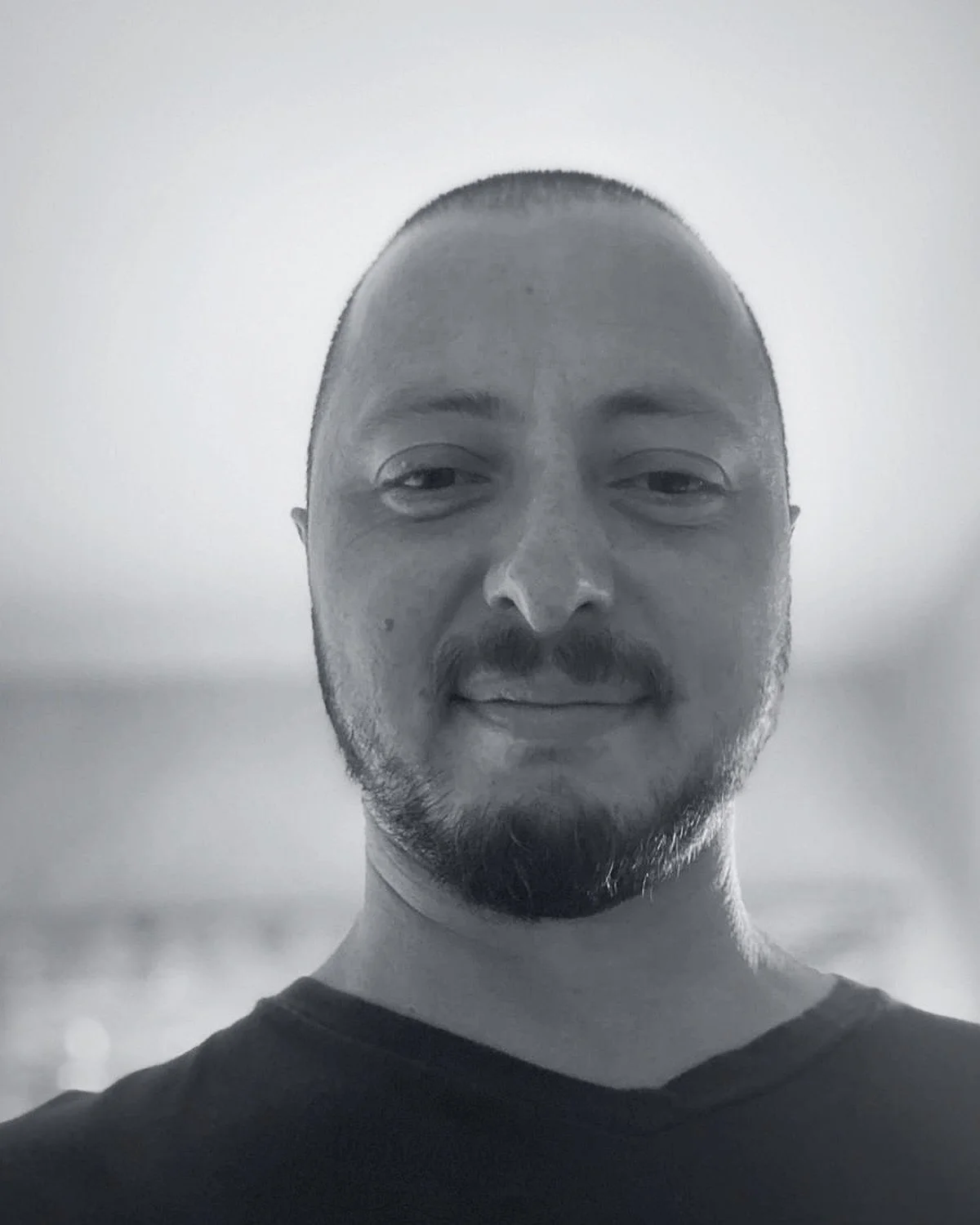 Black and white photo of a man with short hair and a beard, smiling slightly, wearing a dark shirt.