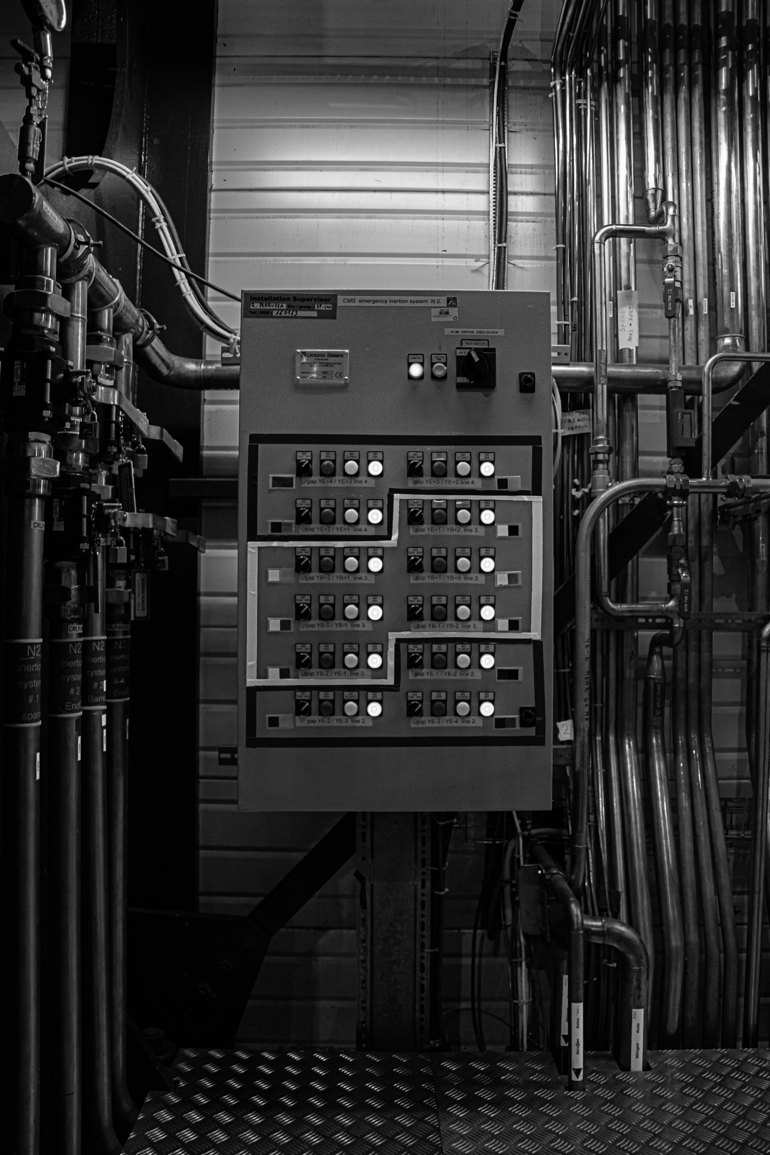 A control panel with switches and labels, surrounded by metallic pipes and fittings in an industrial setting, black and white photo.