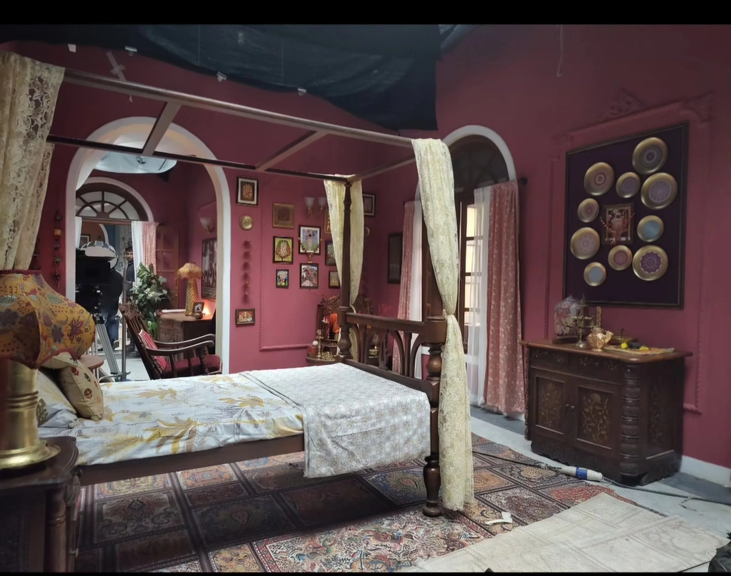 A room with pink walls, decorated with framed pictures and plates. A four-poster bed with floral bedding and cream curtains is in the center. There are windows with drapes, various furniture pieces, and a patterned rug.