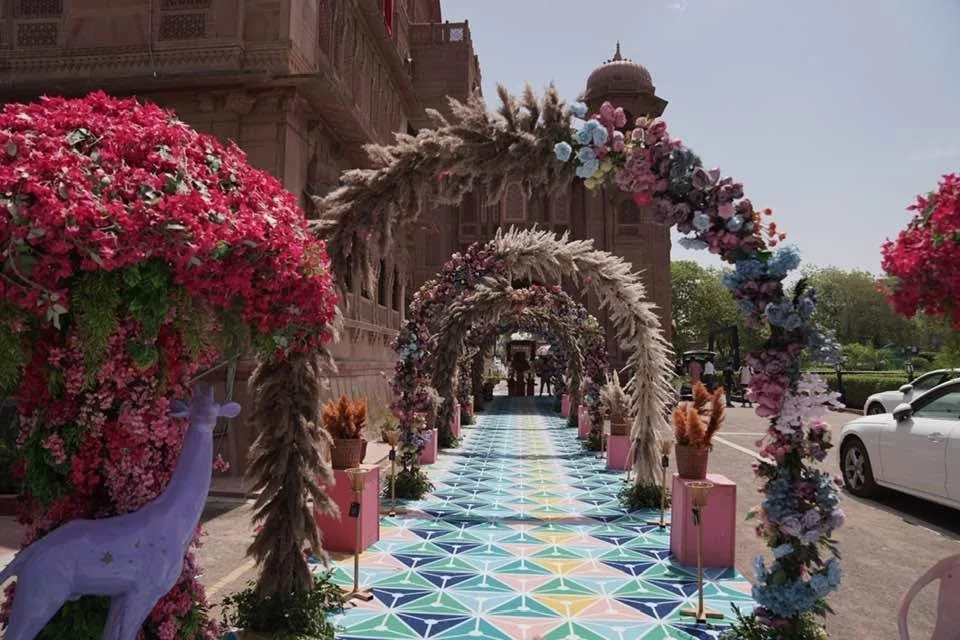 A colorful, decorated walkway with floral arches, pink flower bushes, and potted plants, leading to a grand building with domes and detailed architecture. White cars are parked nearby on the right.