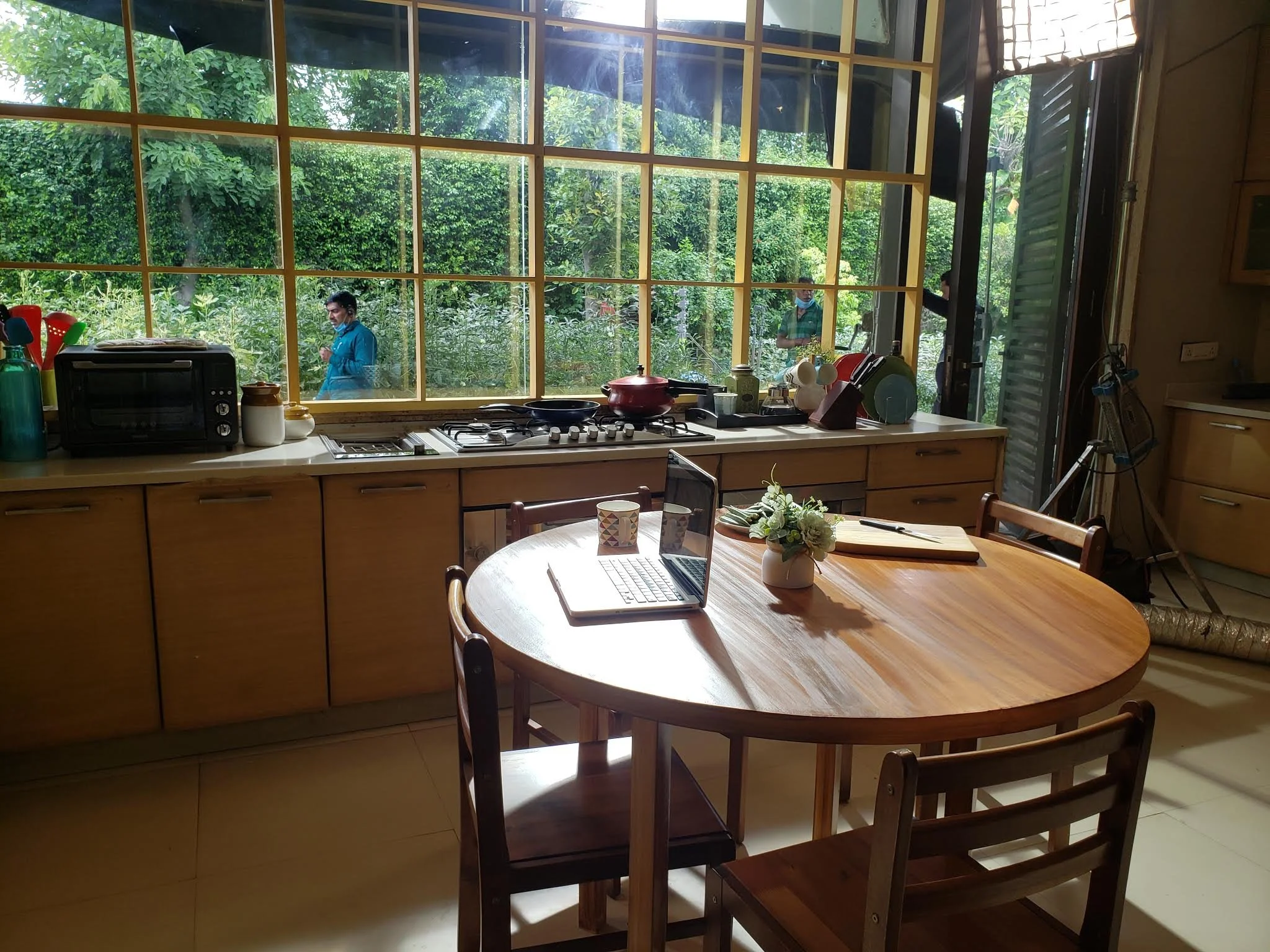 Kitchen interior with a round wooden dining table, a laptop, and a vase with flowers. Large window showing outside greenery with two people, one in a blue shirt and face mask, and another partially visible, outside.