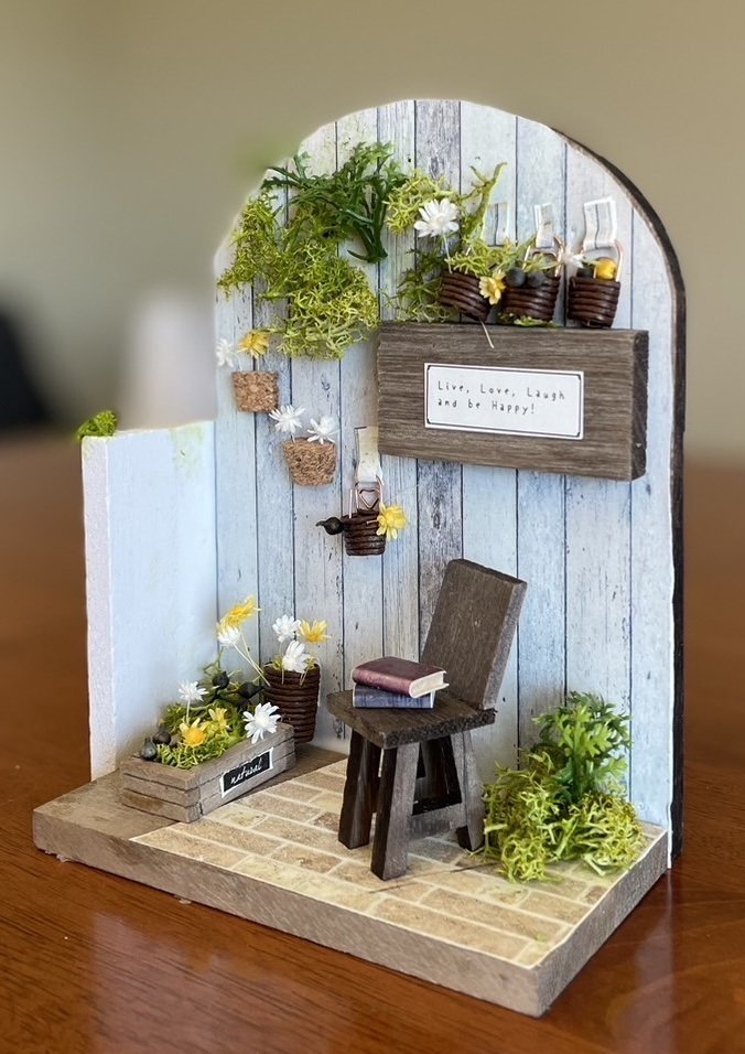 Miniature decorative scene with a wooden chair, books, potted plants, and a sign that reads 'Live, Love, Laugh and be Happy' on a rustic patio backdrop.
