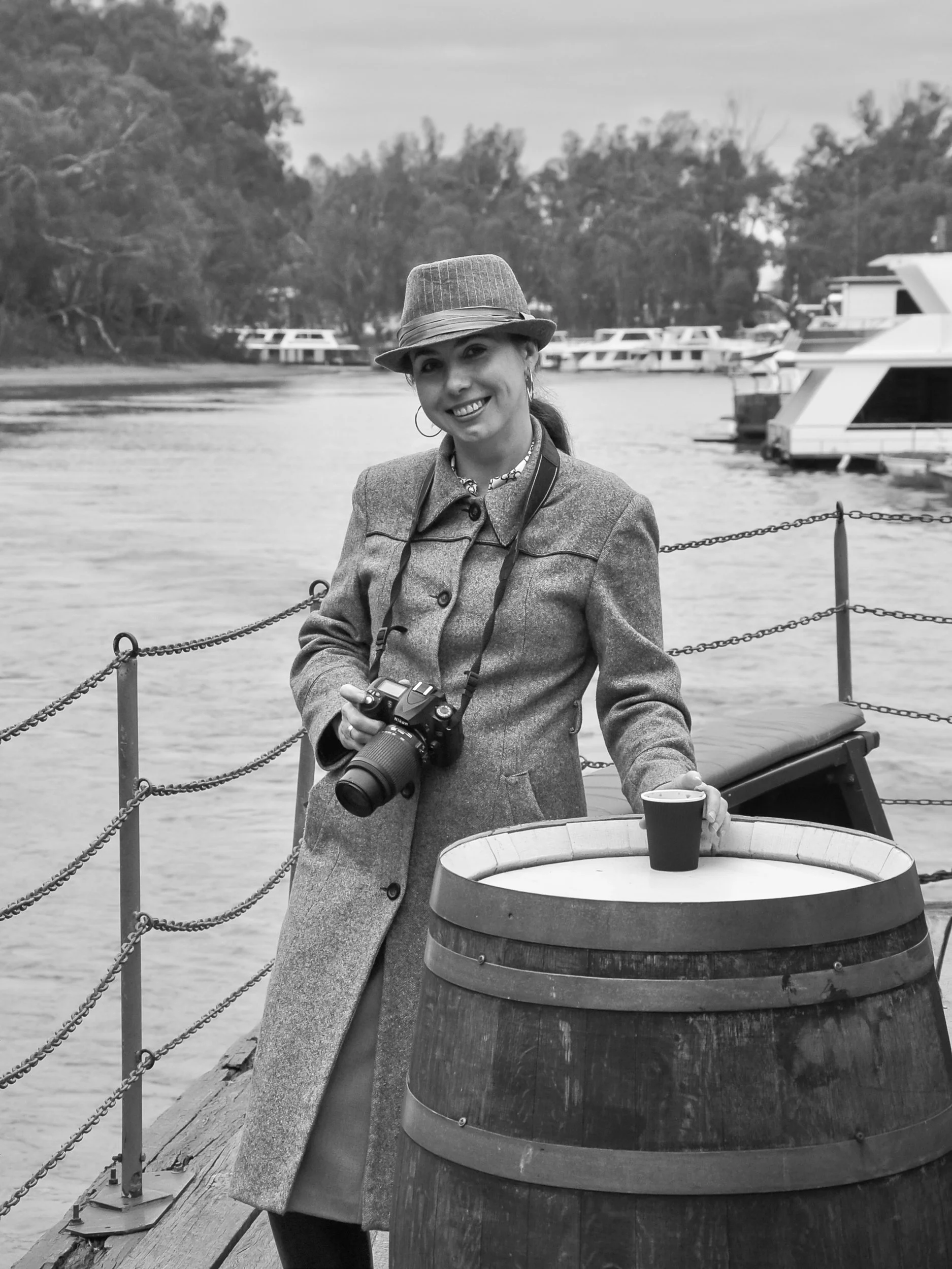 A woman smiling and holding a camera and a cup, standing near a barrell and a railing by a body of water with boats and trees in the background.