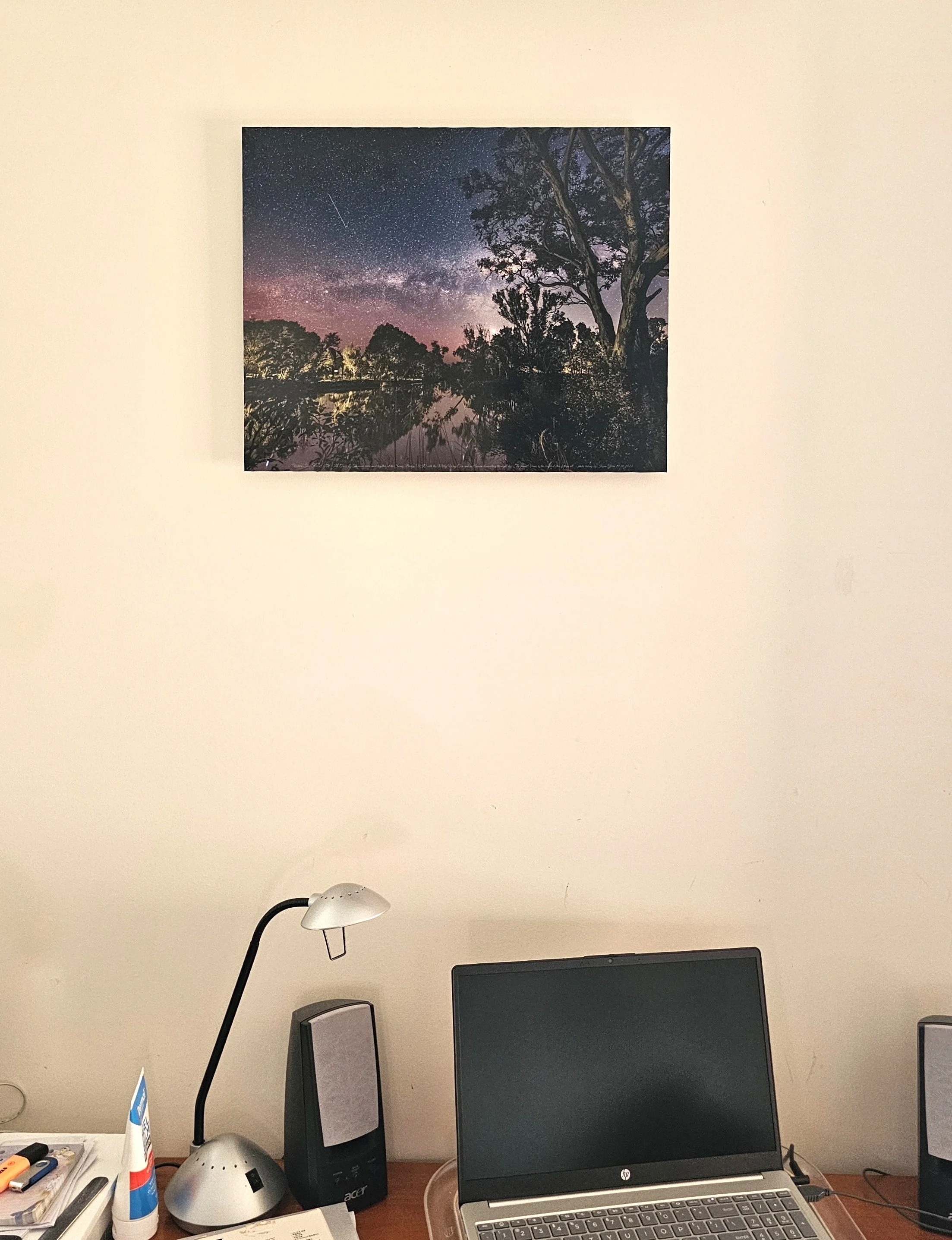 A computer desk setup with a laptop, speakers, a lamp, a tube of glue, papers, and office supplies. There is a wall with a framed picture of a starry night sky above the desk.