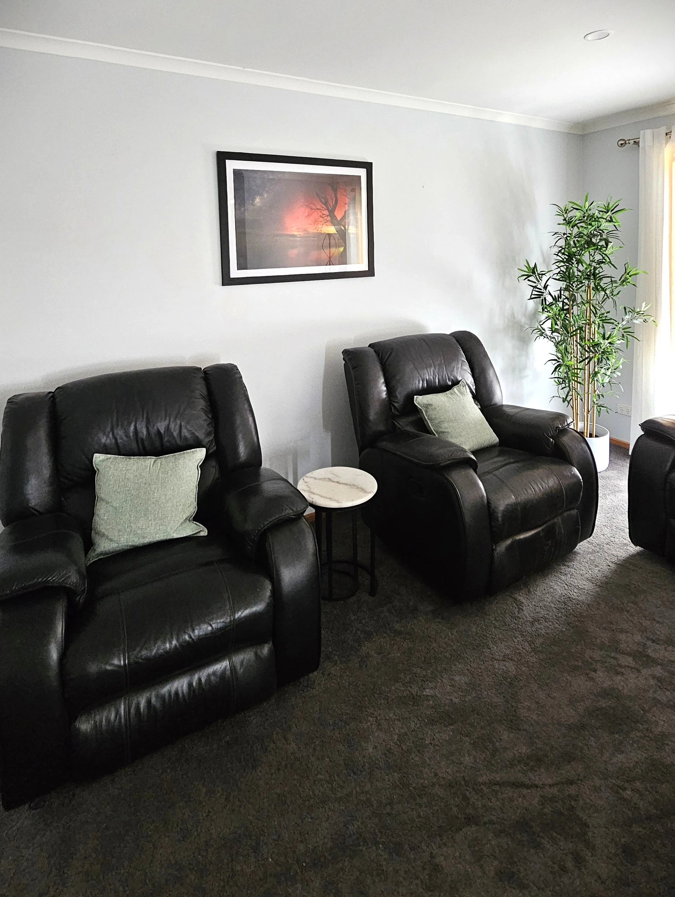 Living room with two black leather recliner chairs, a small round marble side table between them, a green pillow on each chair, a tall potted plant in the corner, a photo of Aurora with a tree on the wall, and a window with white curtains.