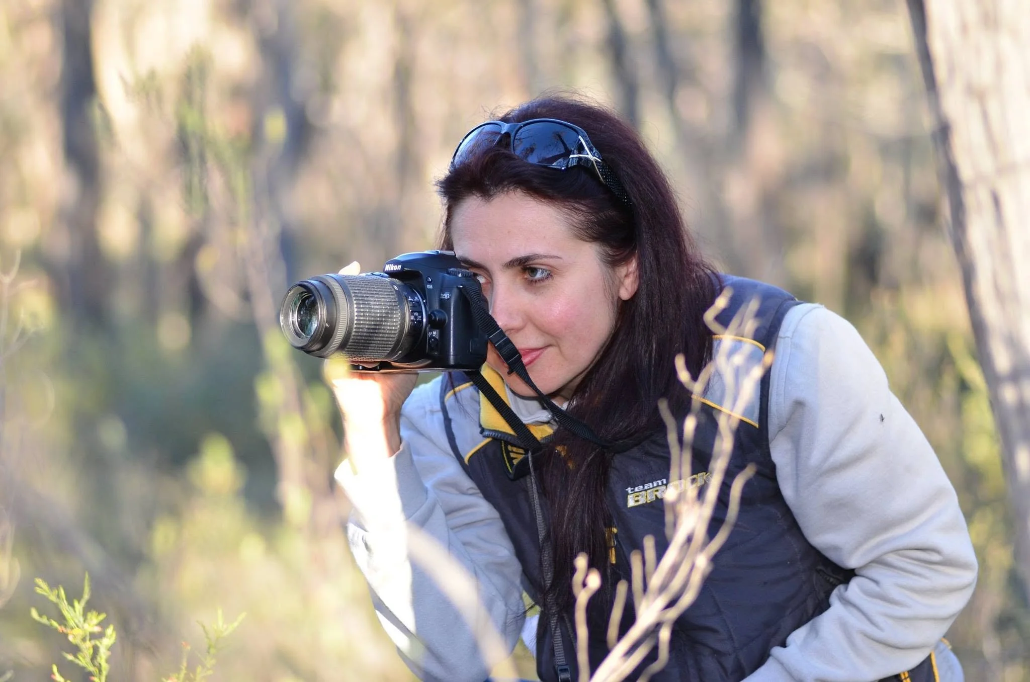 A woman with dark hair and sunglasses on her head is using a Nikon camera to take a picture outdoors in a wooded area during daylight.