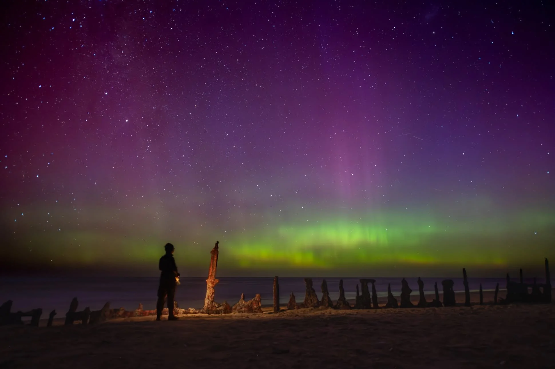 Northern lights in the night sky above a beach with silhouettes of people and wooden posts.
