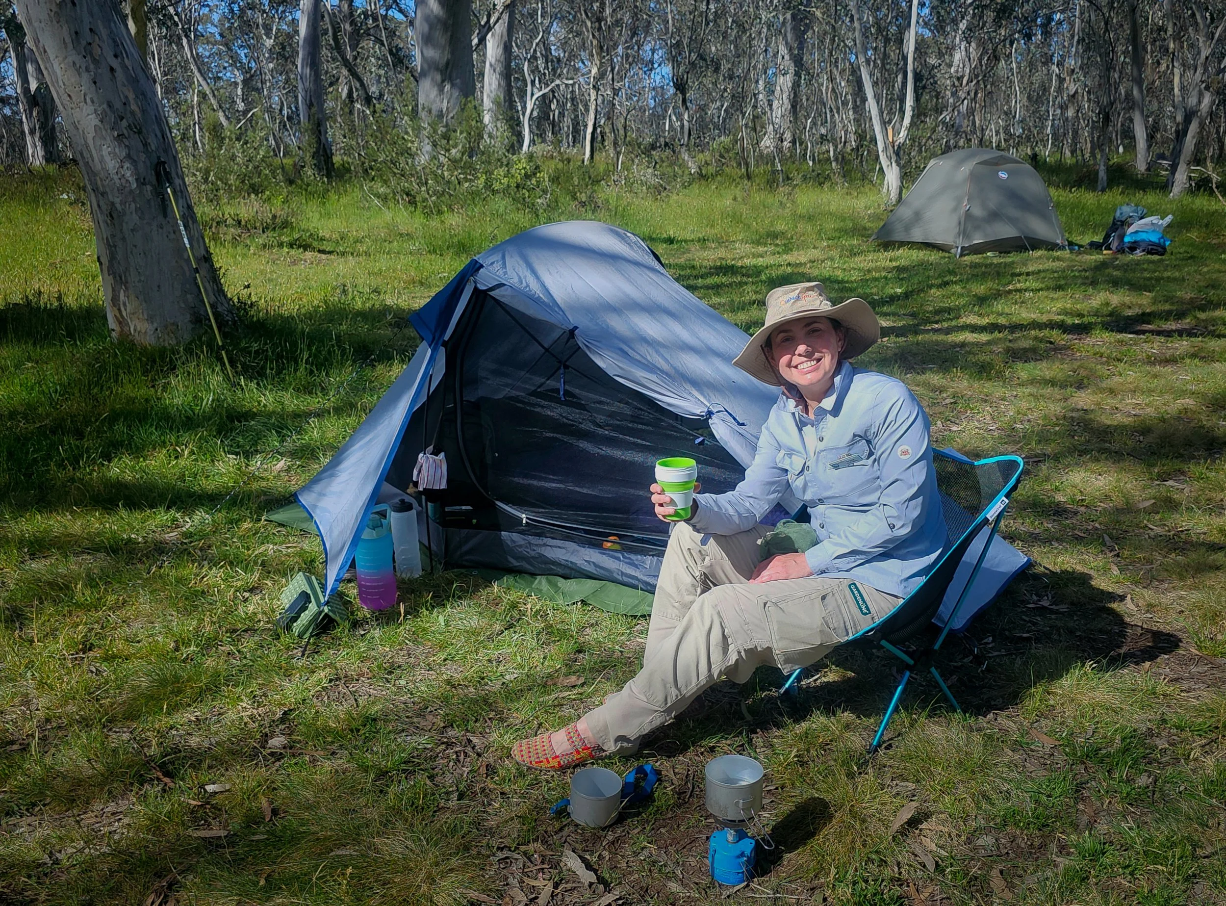 A woman sitting next to a small camping tent in a forested area, holding a green and white cup, with camping gear and other tents visible in the background on a grassy patch.
