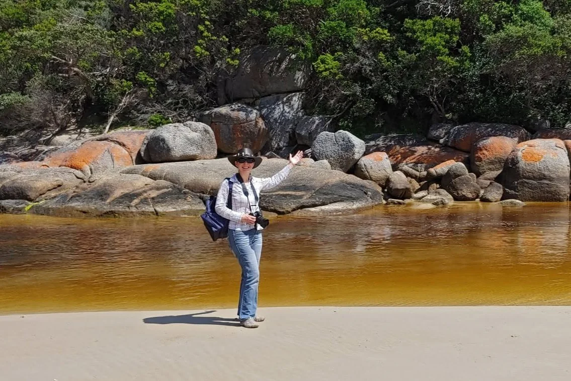 A woman standing on a sandy riverbank with a wooded background, pointing towards large boulders near the water.