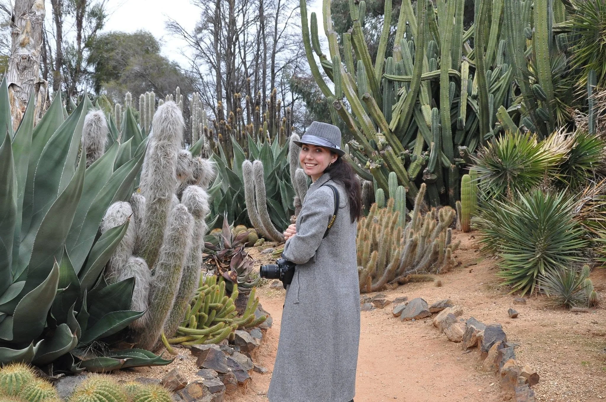 A woman photographer in a gray coat and hat stands in a desert garden among various tall cacti and succulents, smiling at the camera.