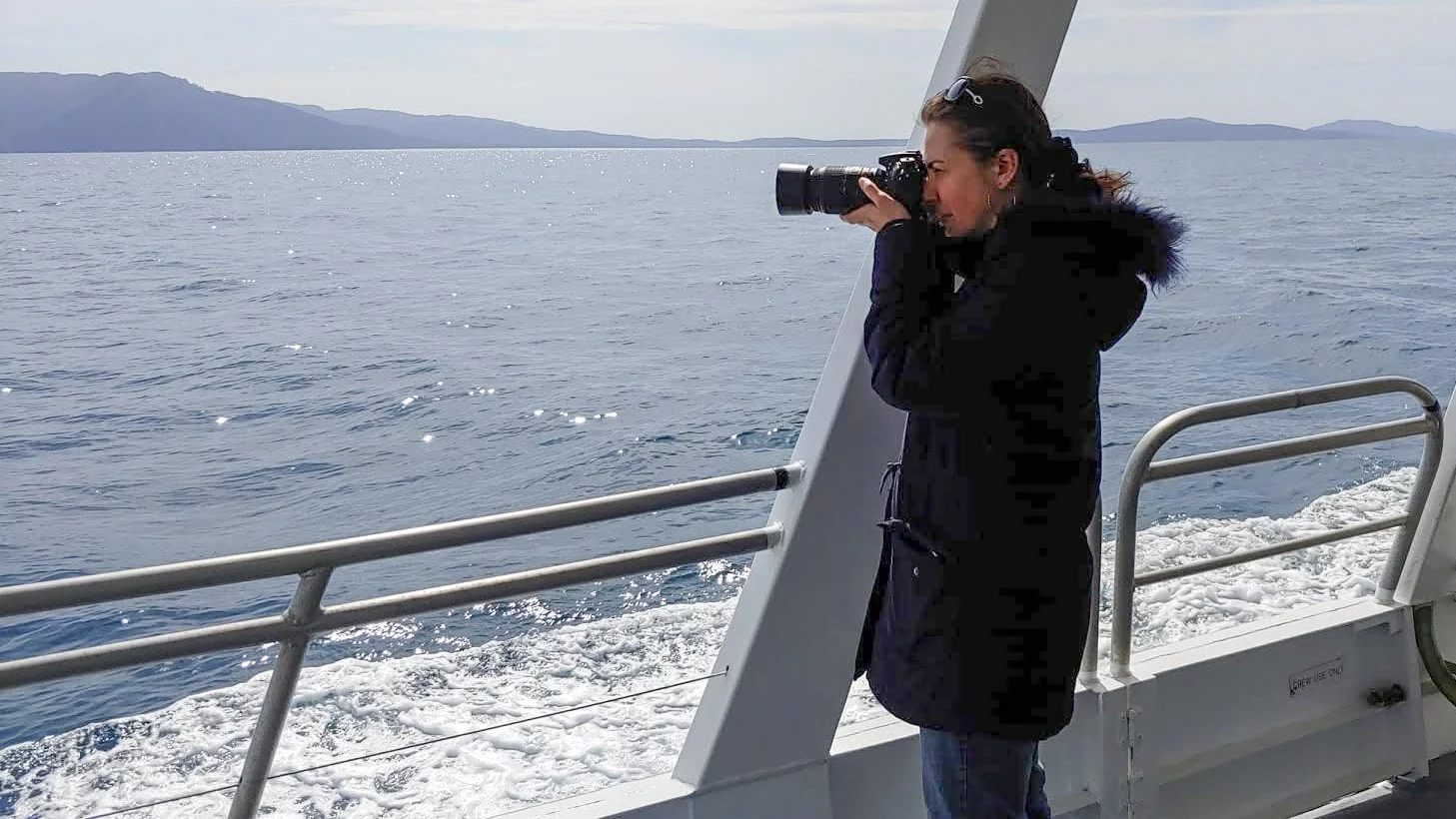 A woman taking photographs with a camera on a boat in the ocean, with distant mountains in the background.