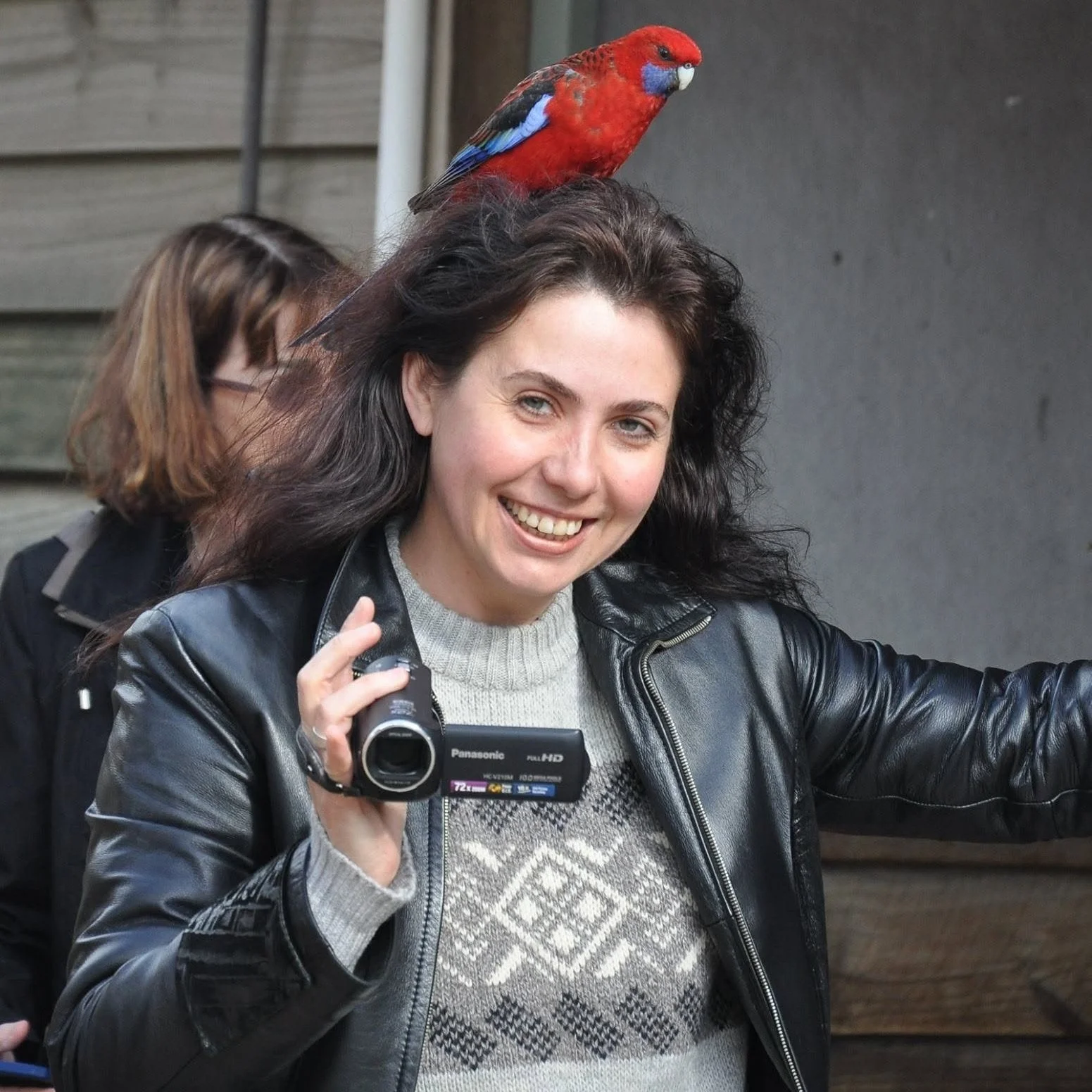 A woman with brown hair and green eyes smiling outdoors while holding a camera, with a colorful red and blue bird perched on her head.