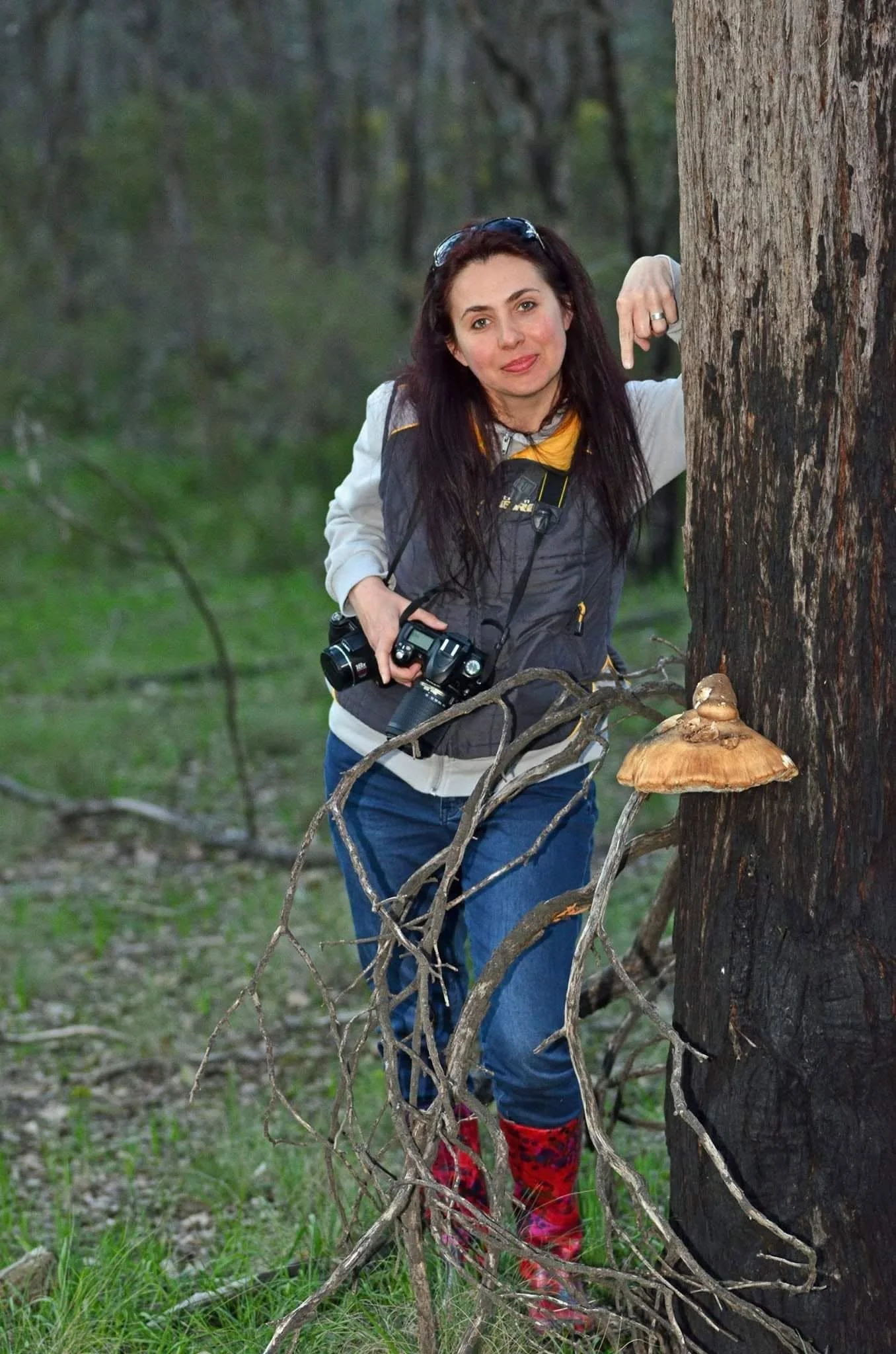 A woman with dark hair, sunglasses on her head, standing behind a large tree with roots, in a forest. She is smiling, wearing a gray jacket, a camera around her neck, blue jeans, and red patterned boots.