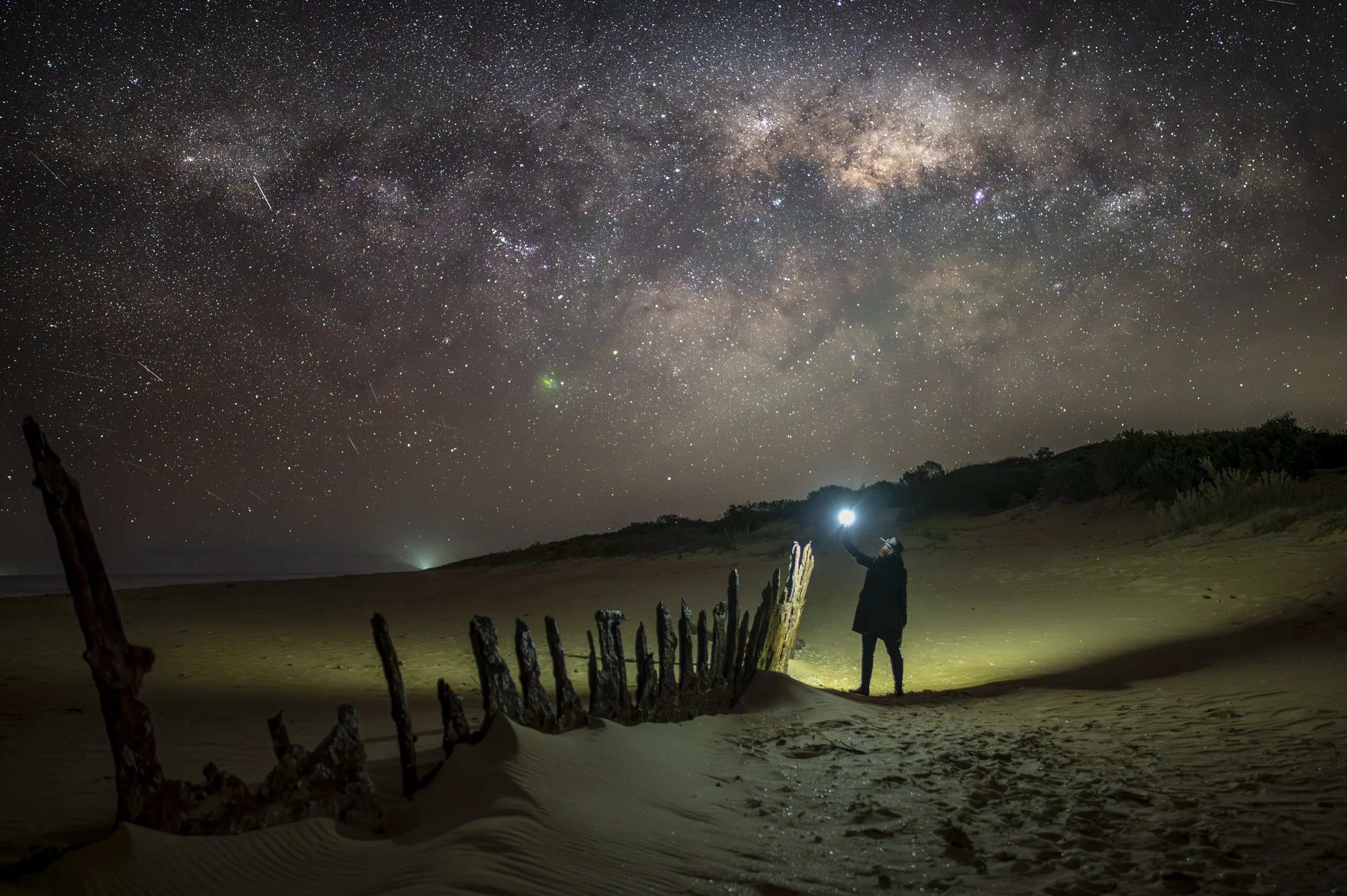 Person in dark clothing holding a light on a sandy beach at night, looking up at a star-filled sky with the Milky Way galaxy.