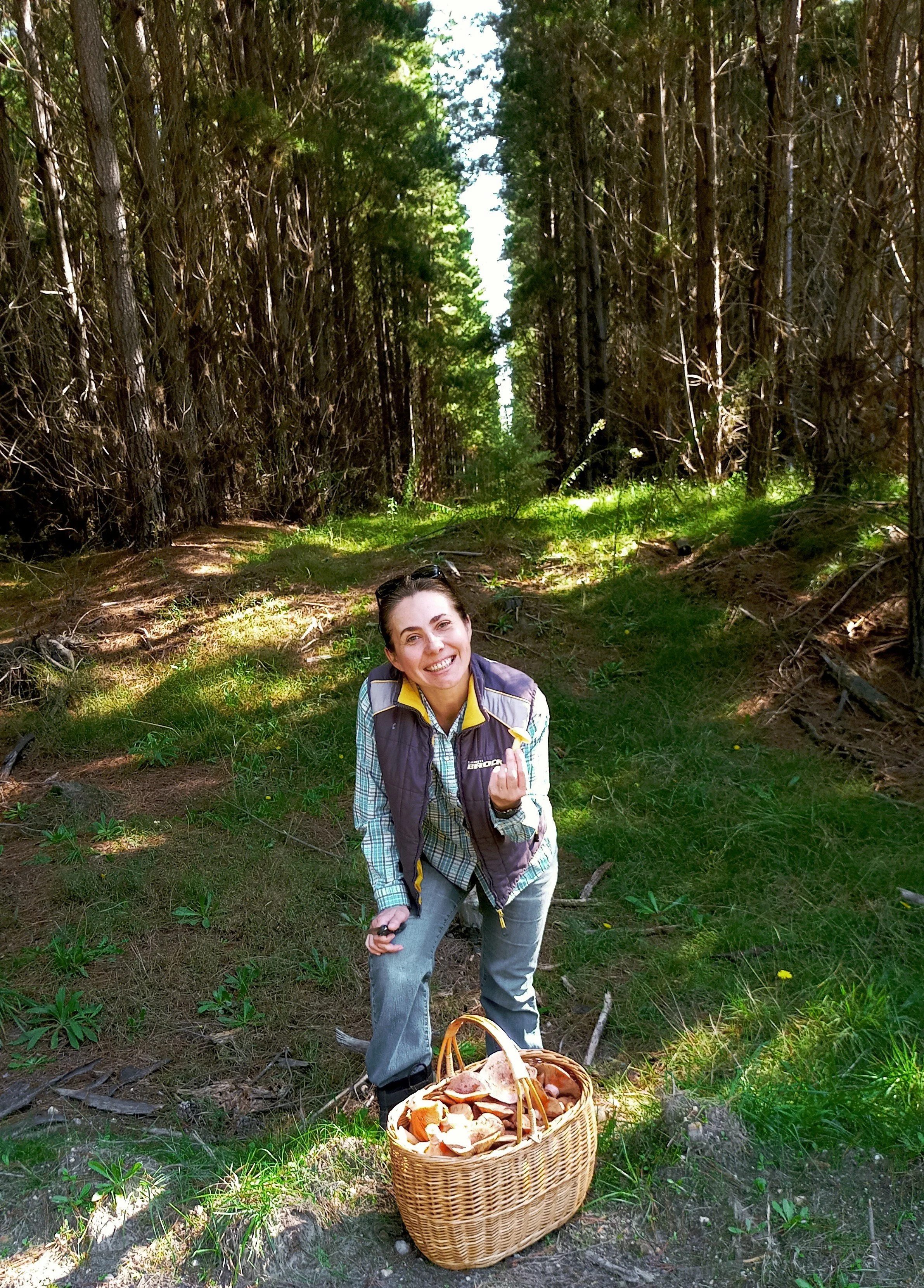 A woman smiling in a forest with a basket of freshly picked mushrooms on the ground in front of her.