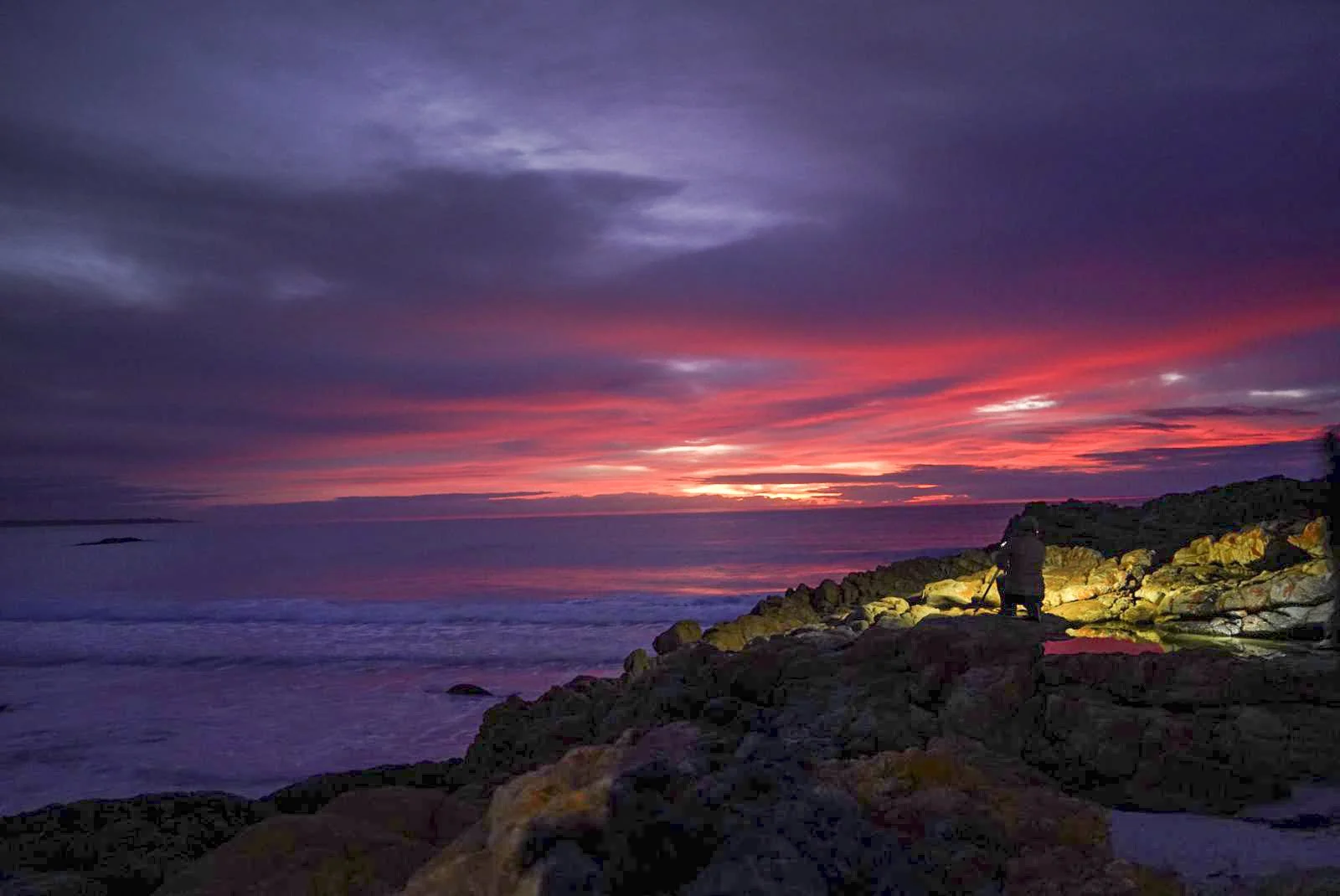 Person photographing a colorful sunset over the ocean from a rocky shoreline.