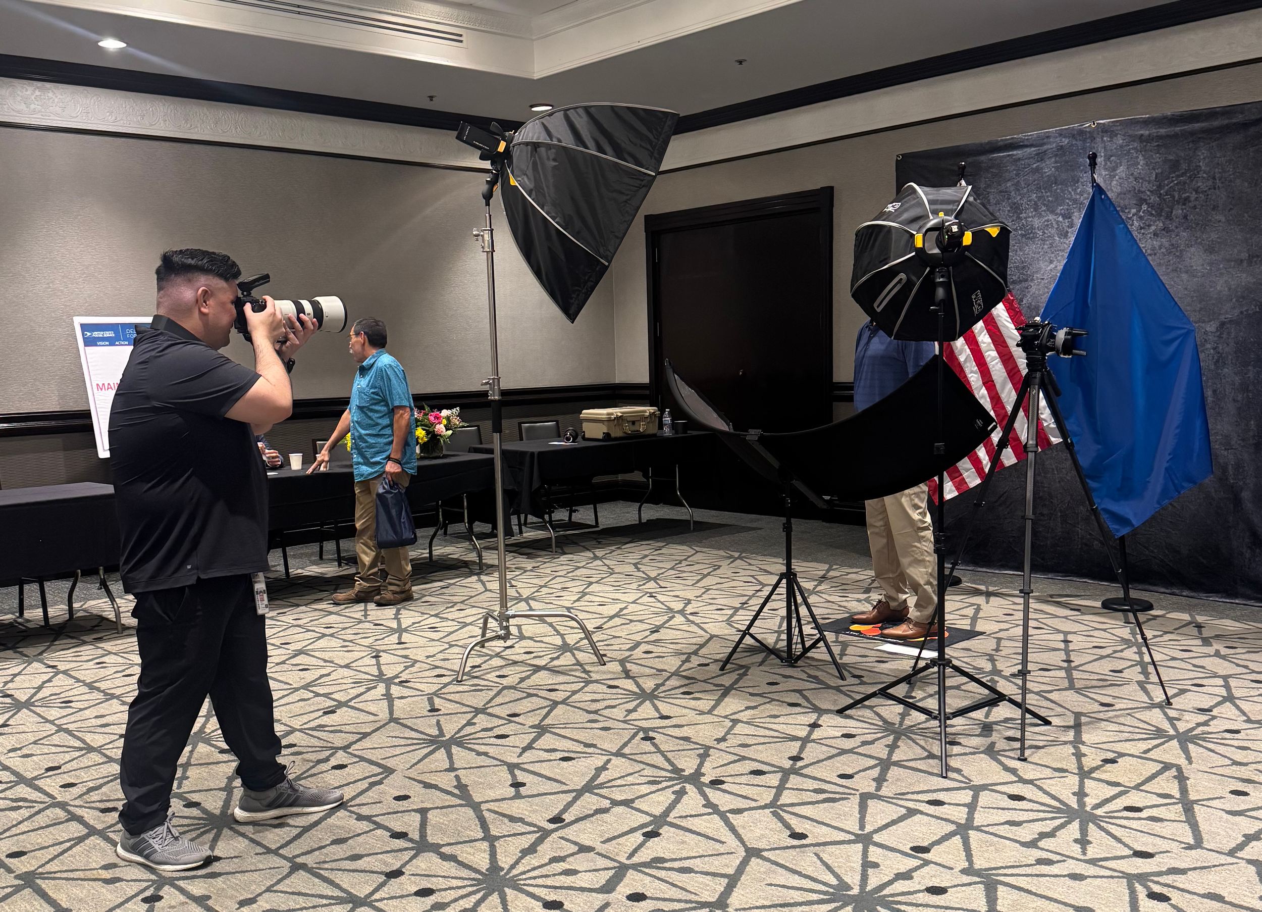 A man with a camera taking a photograph of a person standing near a table with flowers, in a conference room with studio lighting equipment set up.