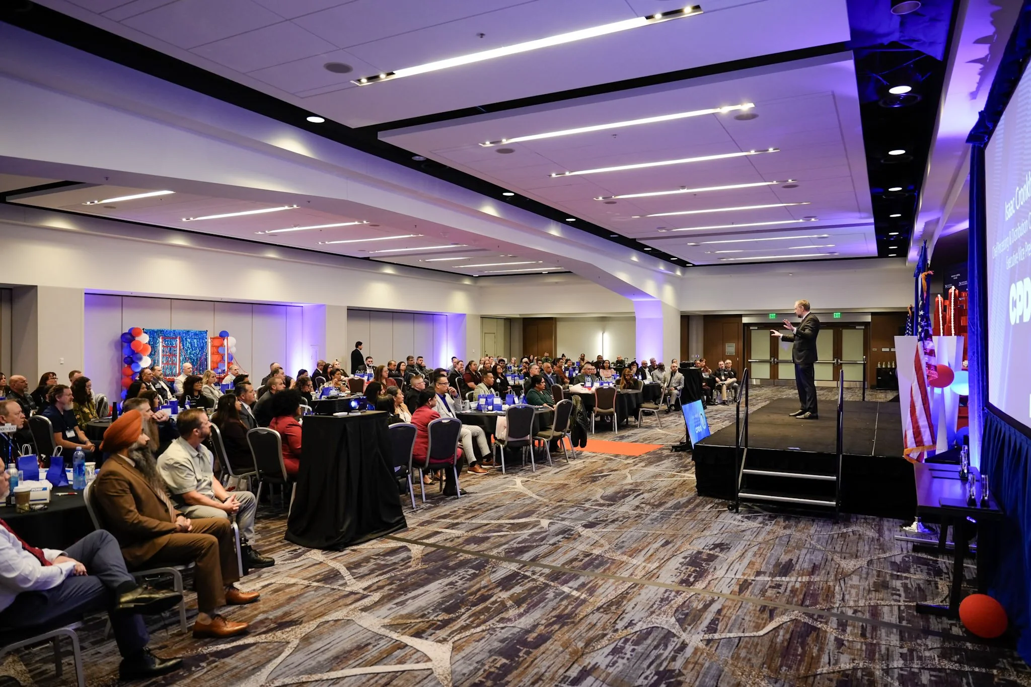 A conference room filled with attendees seated at tables and listening to a speaker on stage. The room is decorated with balloons and patriotic symbols, and a large screen displays a presentation.