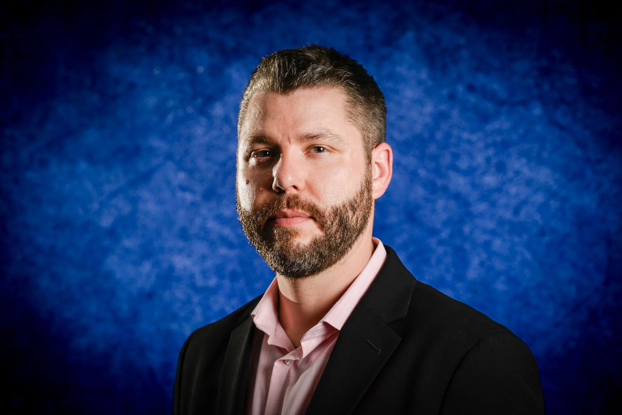 Portrait of a man with short dark hair, beard, wearing a black suit and pink shirt, against a blue textured background.