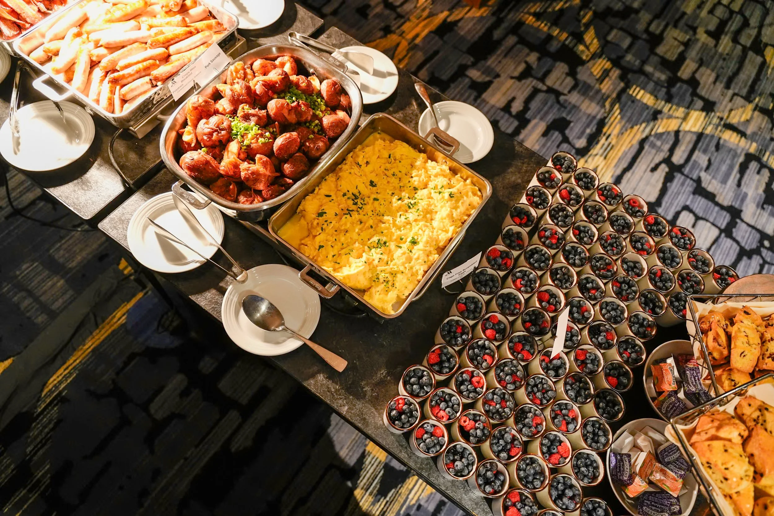 Buffet table with trays of breakfast foods, including scrambled eggs, breakfast sausage meatballs with chopped green onions, and crab meat sticks, alongside small cups filled with blueberries and strawberries topped with out-of-focus blackberries and