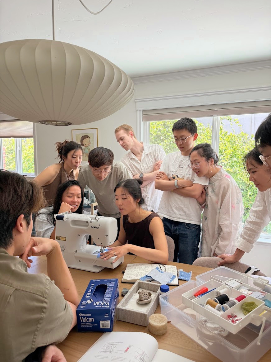 A group of people gathered around a woman sewing on a sewing machine at a table in a bright room with large windows.