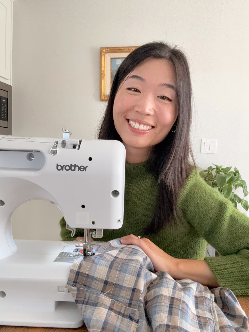 A woman smiling at a sewing machine with fabric, in a room with a framed picture and houseplants.