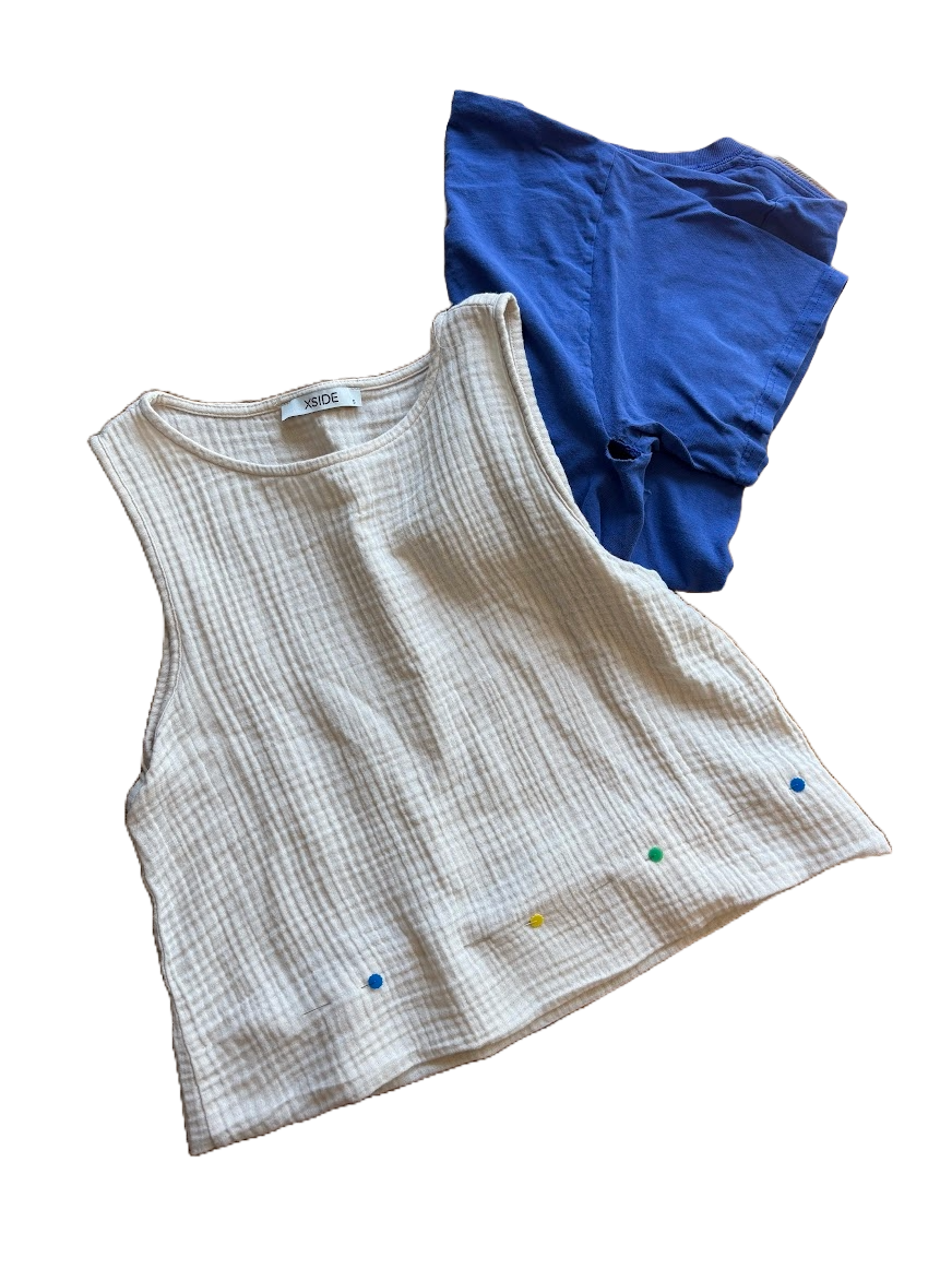A beige sleeveless top with vertical ribbing and three pins on the bottom, next to a blue T-shirt with a hole in it.