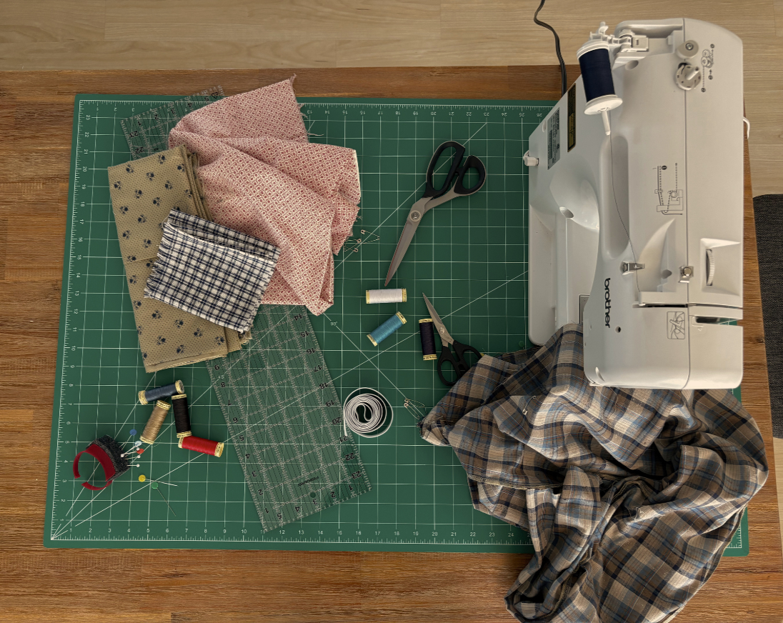 A sewing workspace with fabric, scissors, thread, pins, ruler, and a sewing machine on a green cutting mat on a wooden table.