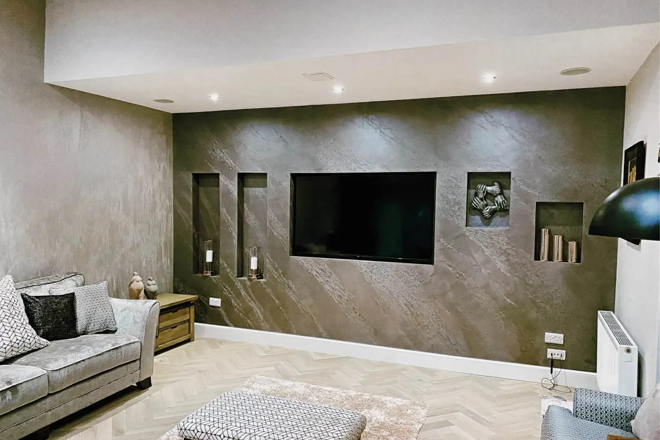 Modern living room with a textured dark accent wall, mounted flat-screen TV, built-in wall niches with decorative items, a light-colored sofa with patterned cushions, a wooden side table, a small gray ottoman, and a wall-mounted lamp.