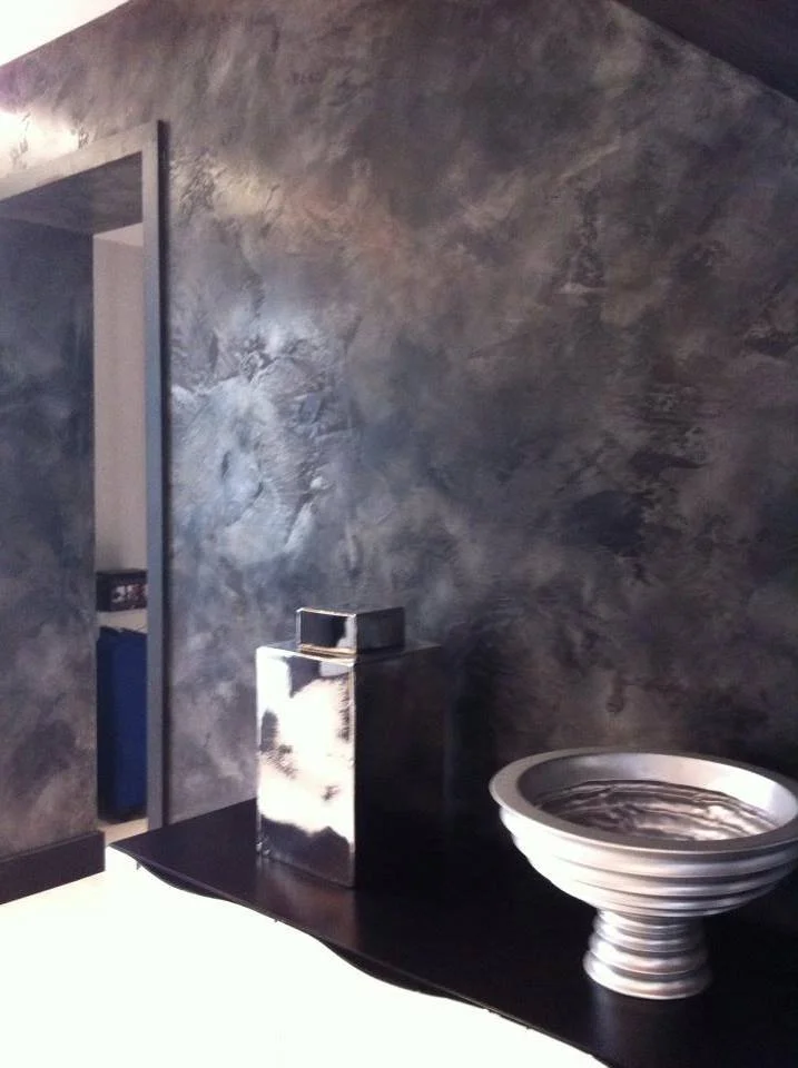A bathroom with dark, textured walls, a modern square sink on a black countertop, a tall reflective metallic container, and a round, ceramic vessel sink.