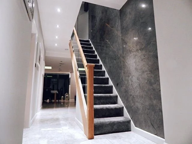 Interior view of a home with a staircase covered in dark carpet, white walls, and a marble accent wall on the right side that's made of Venetian plaster.