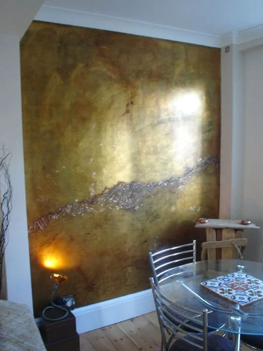 Interior of a room with a large, textured gold-colored accent wall, a glass dining table with metal chairs, and a small shelf on the ceiling. There is a small lamp on a stand on the left side and some decorative items on the shelf.