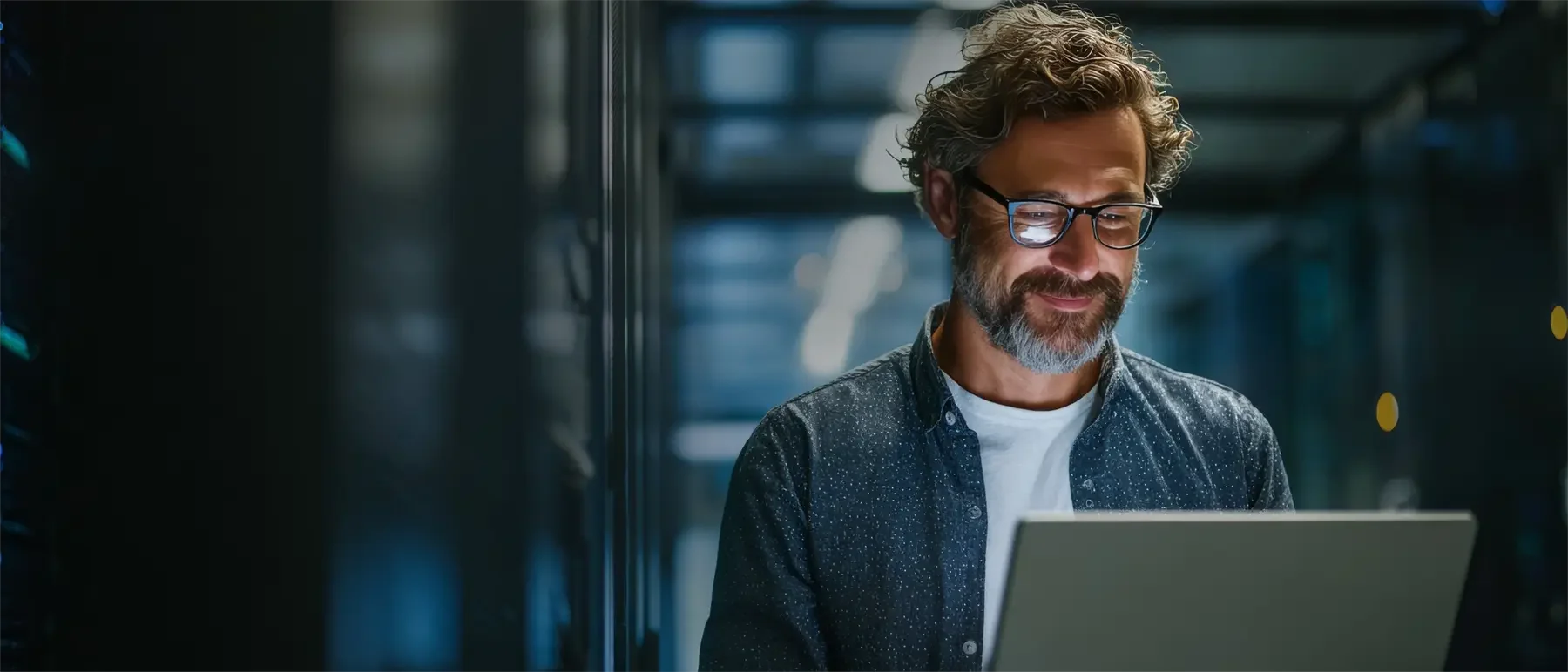 Ein Mann mit Brille und grauem Bart arbeitet in einem Serverraum an einem Laptop.