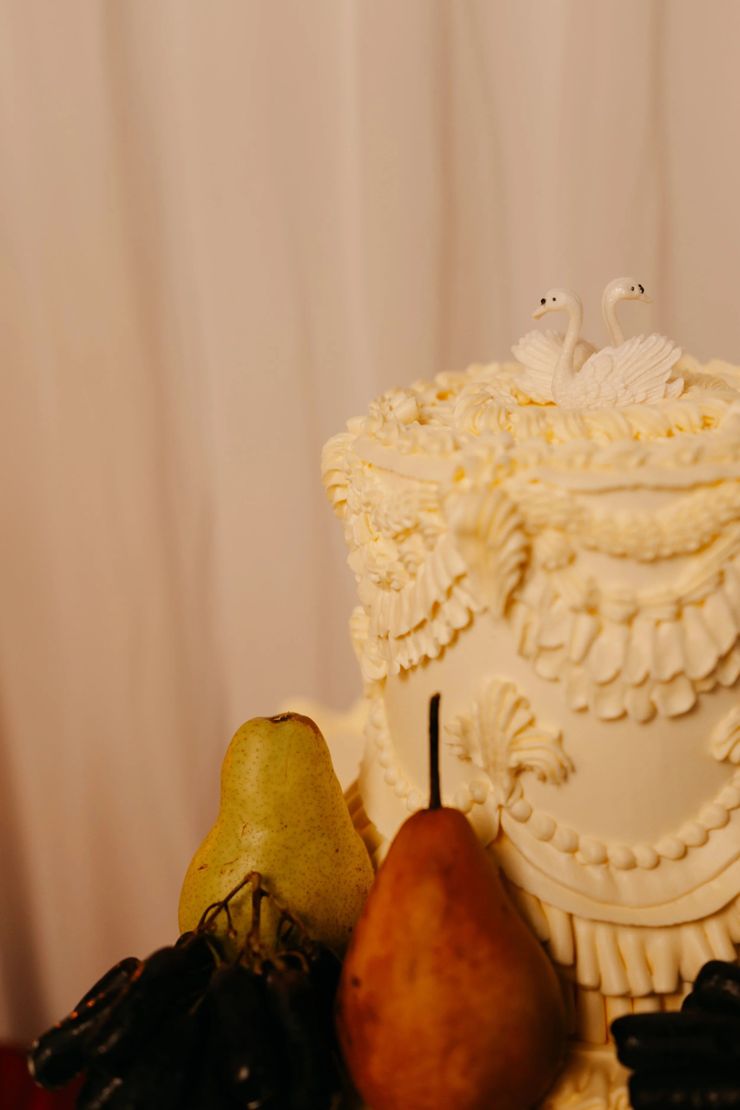 An ornate white cake decorated with swan-shaped toppers and piped frosting, surrounded by pears, grapes, and apples.