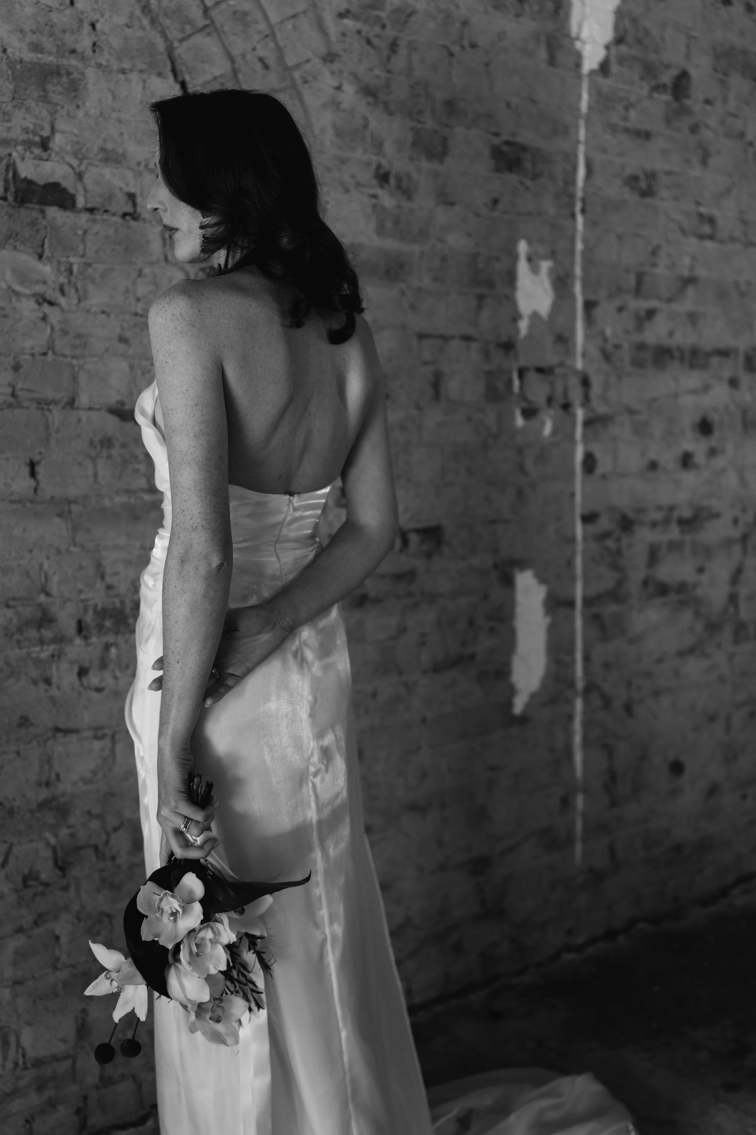 An elegantwoman in a wedding dress holding a bouquet of flowers stands against a brick wall, her face turned to the side, with dark hair in loose waves. Elegant Adelaide South Australian elopement, luxury photography.