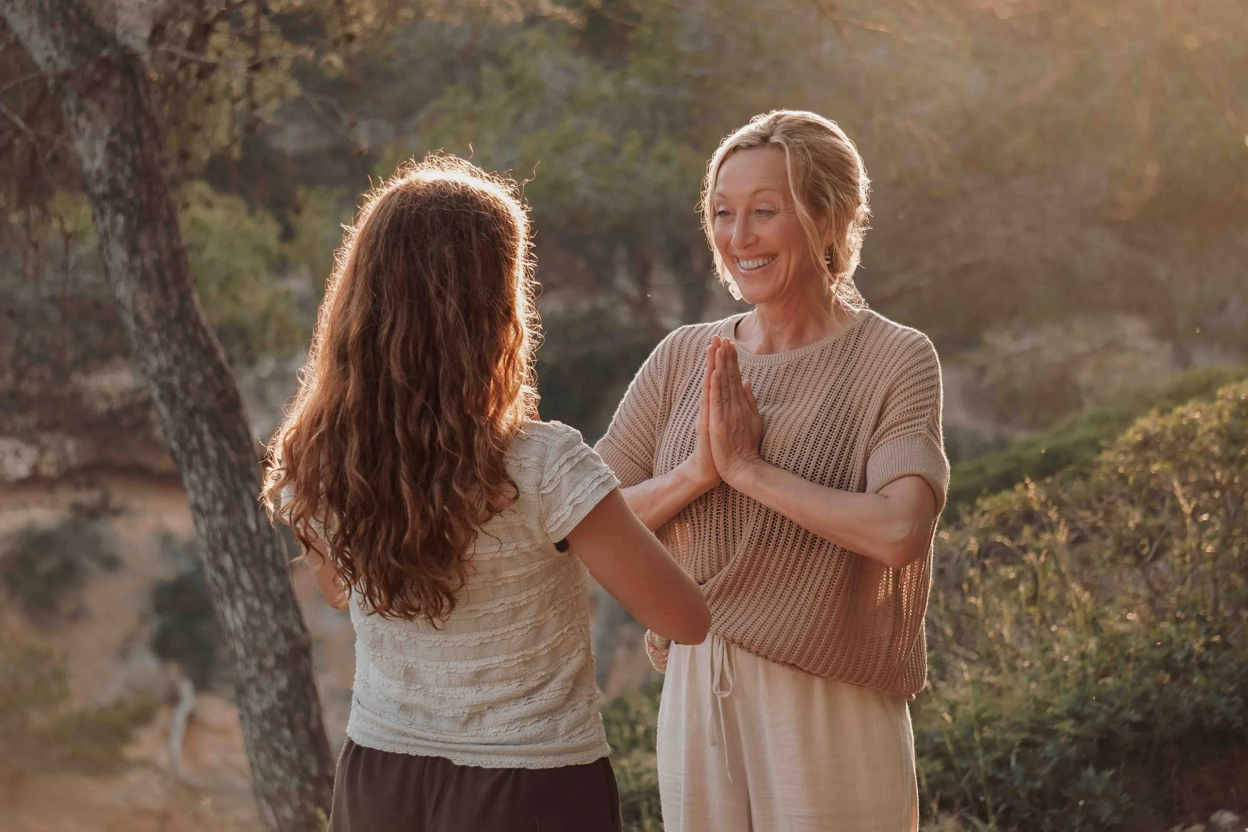 Zwei Frauen stehen sich gegenüber, während sie im Freien bei Sonnenuntergang lächelnd miteinander sprechen. Eine Frau hat lange, lockige braune Haare, die andere kurze, blonde Haare. Beide tragen lockere, helle Kleidung.