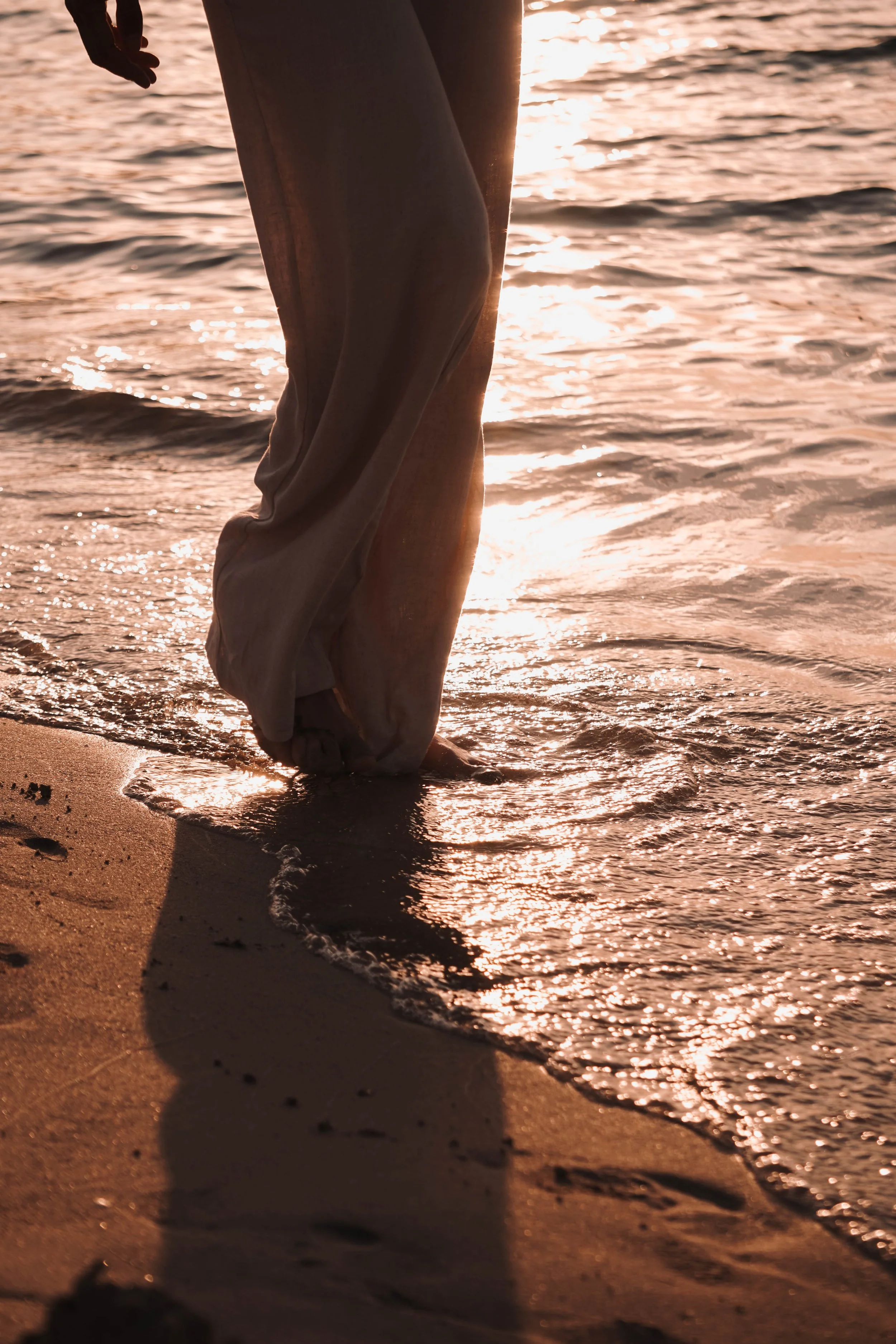 Ein Teil eines menschlichen Beins und Fuß im Sand, mit Blick auf das Meer und den Sonnenuntergang im Hintergrund.