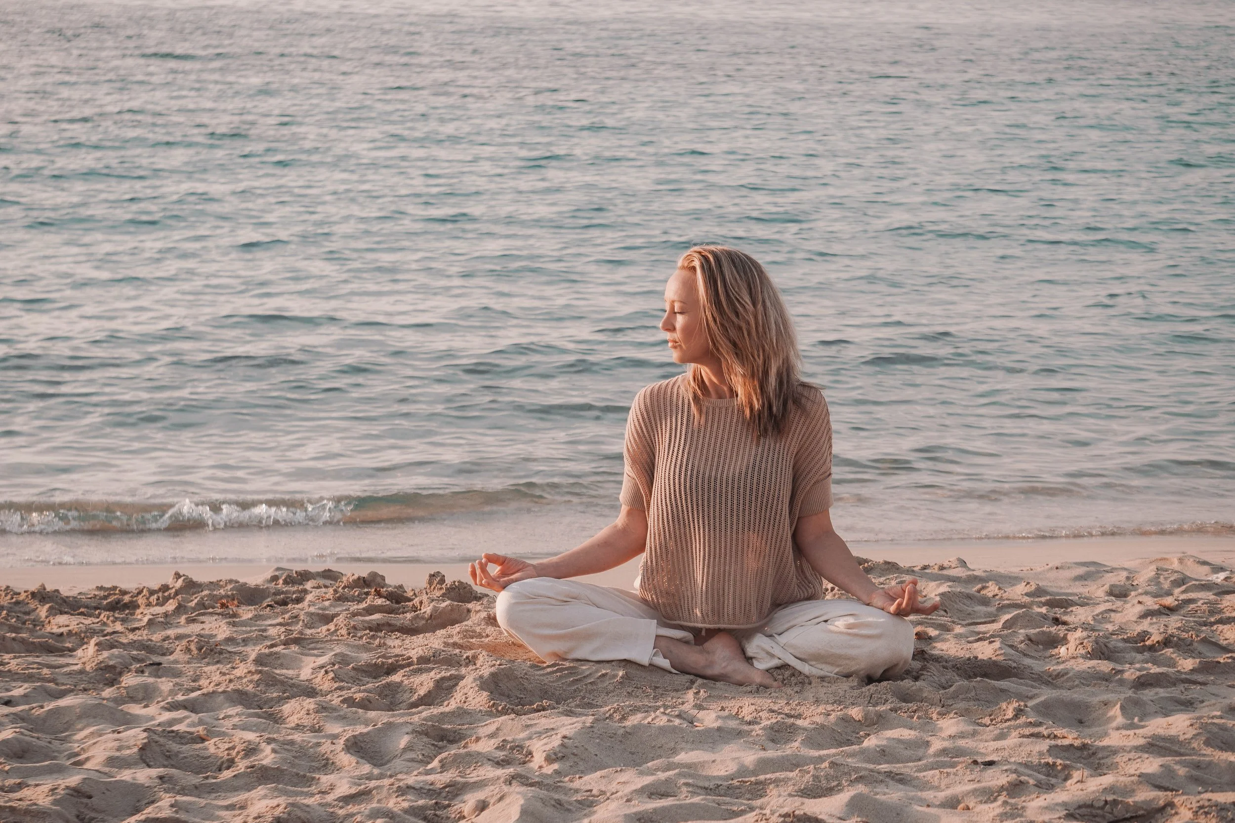 Eine Frau sitzt im Schneidersitz am Strand und meditiert, mit Blick auf das Meer, bei Sonnenuntergang.