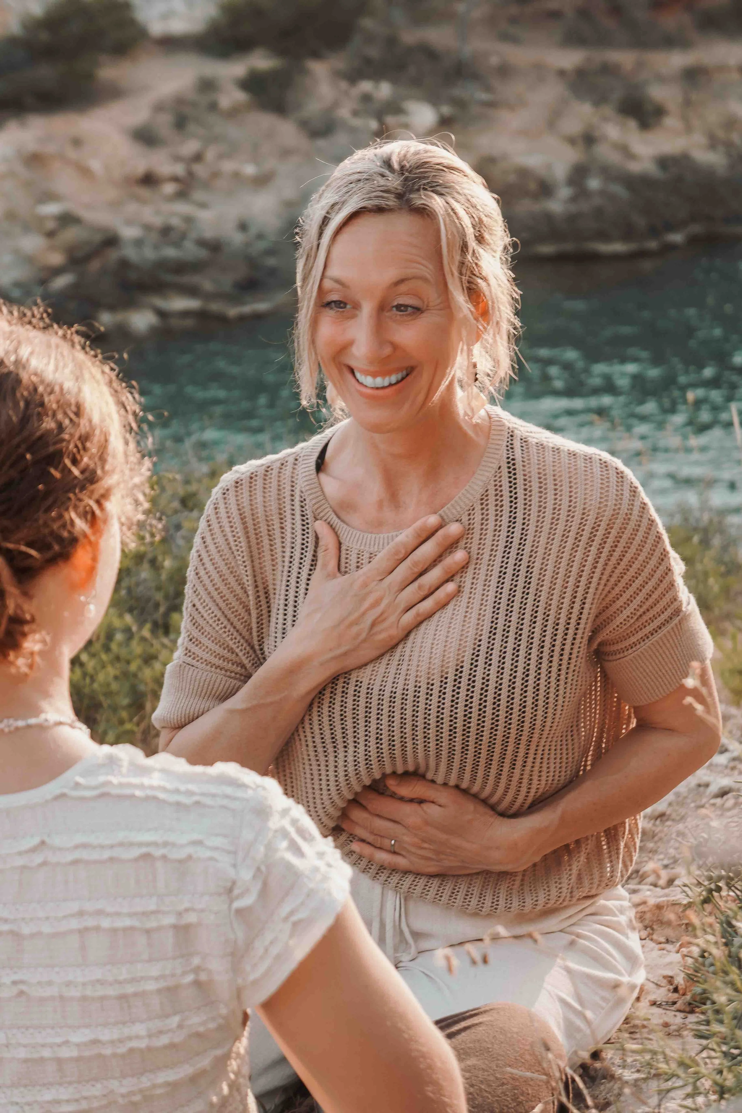Zwei Frauen im Gespräch am Strand, eine Frau berührt die Brust der anderen, beide wirken glücklich und friendly, im Hintergrund Wasser und Felsen.