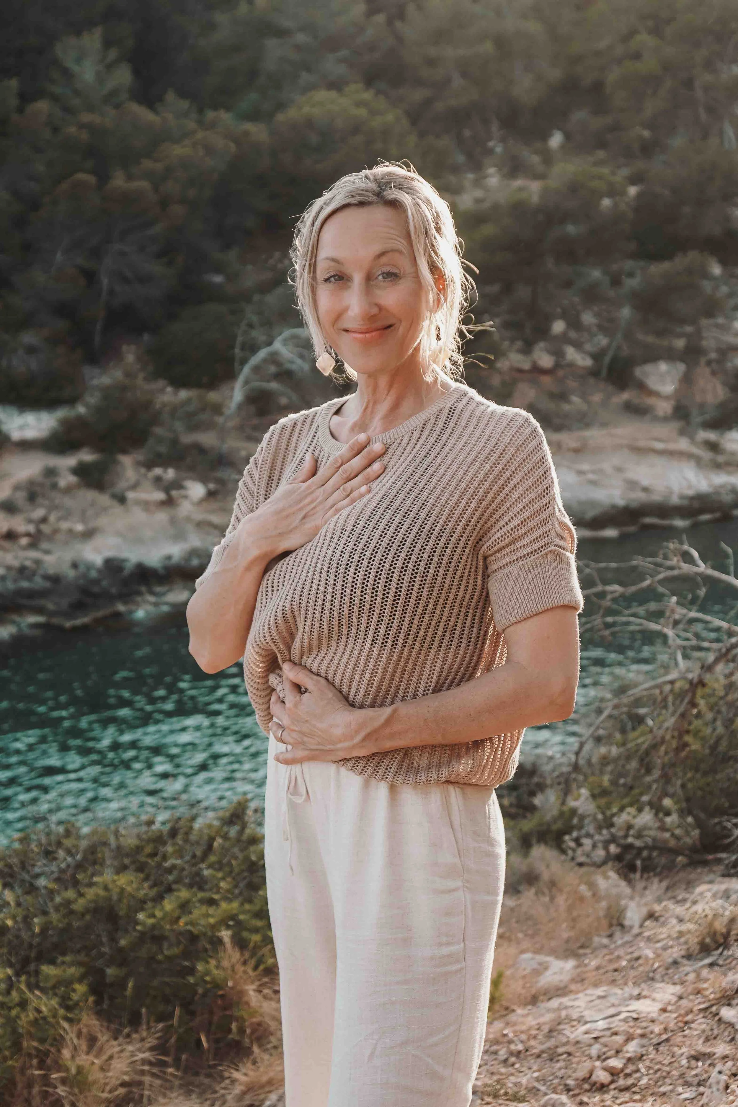 Eine Frau steht an einem Flussufer vor einem bewaldeten Hang, trägt eine beige Strickjacke und ein weißes Kleid, lächelt sanft und legt eine Hand auf die Brust.