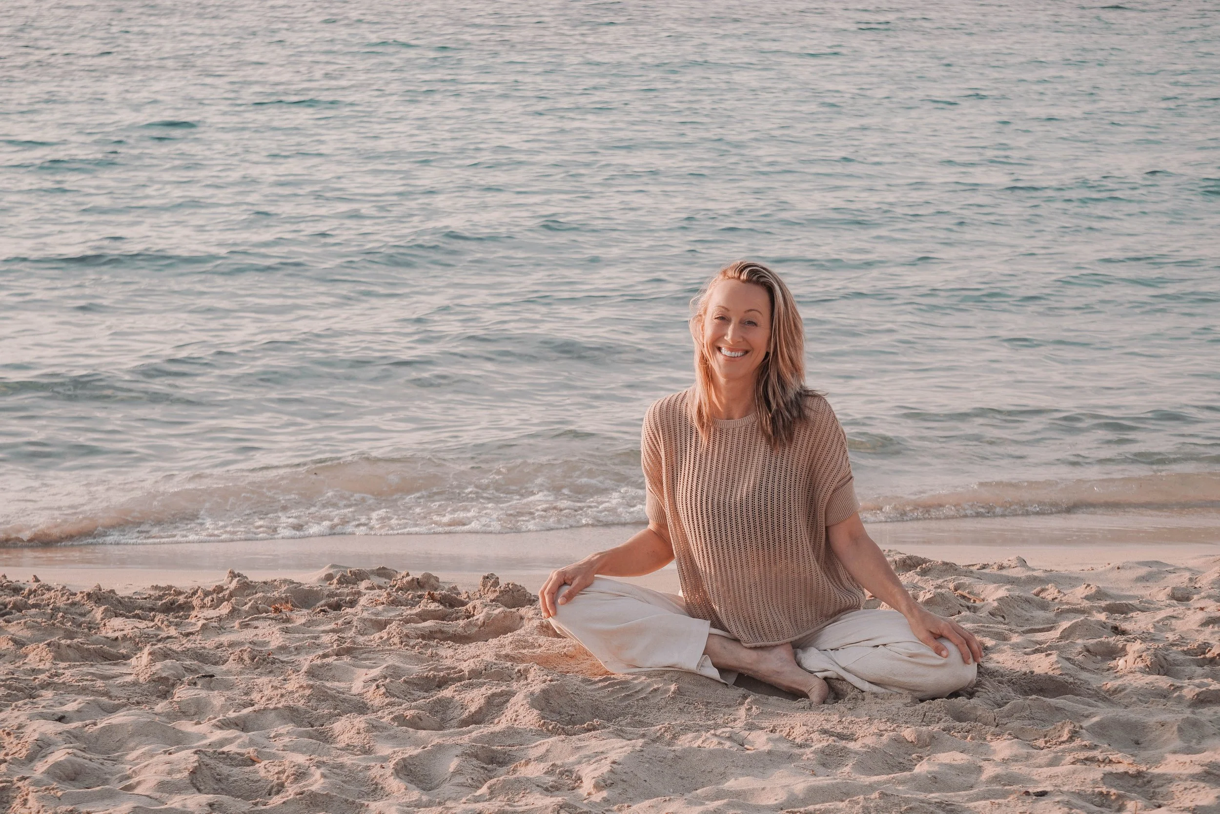Eine lachende Frau sitzt im Sand am Strand mit dem Meer im Hintergrund.