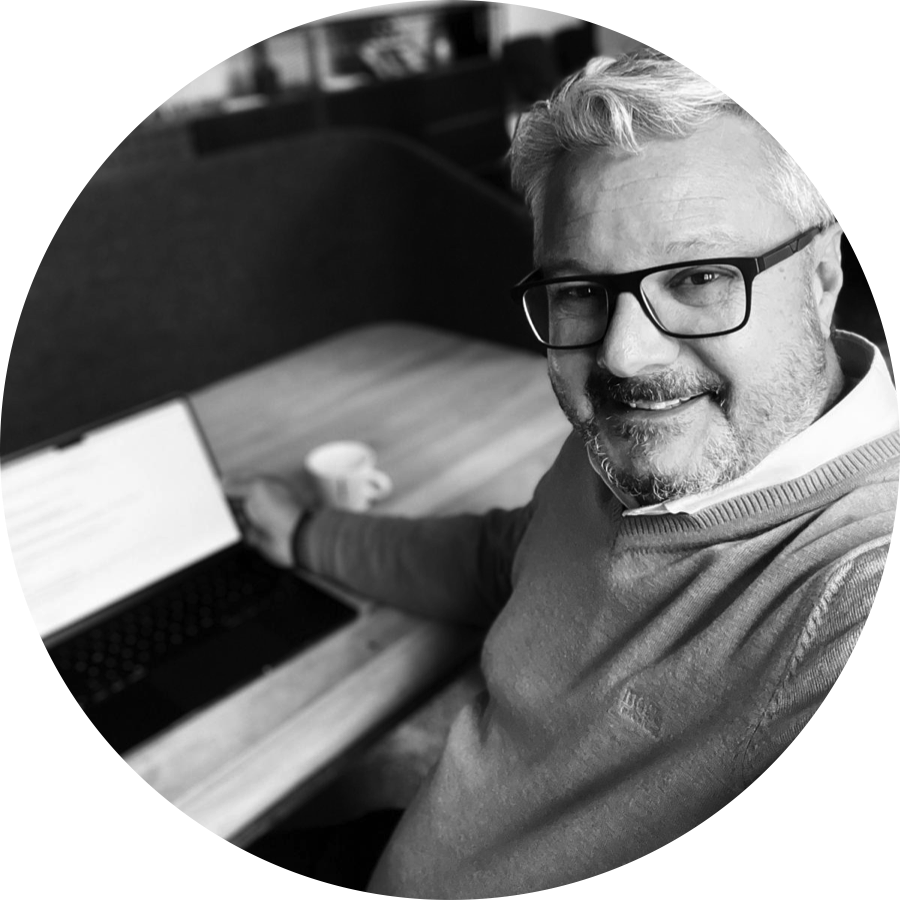 Ein lachender Mann mit Brille sitzt an einem Tisch mit einem Laptop und einer Kaffeetasse, in einem Büro. Schwarz-Weiß-Fotografie.