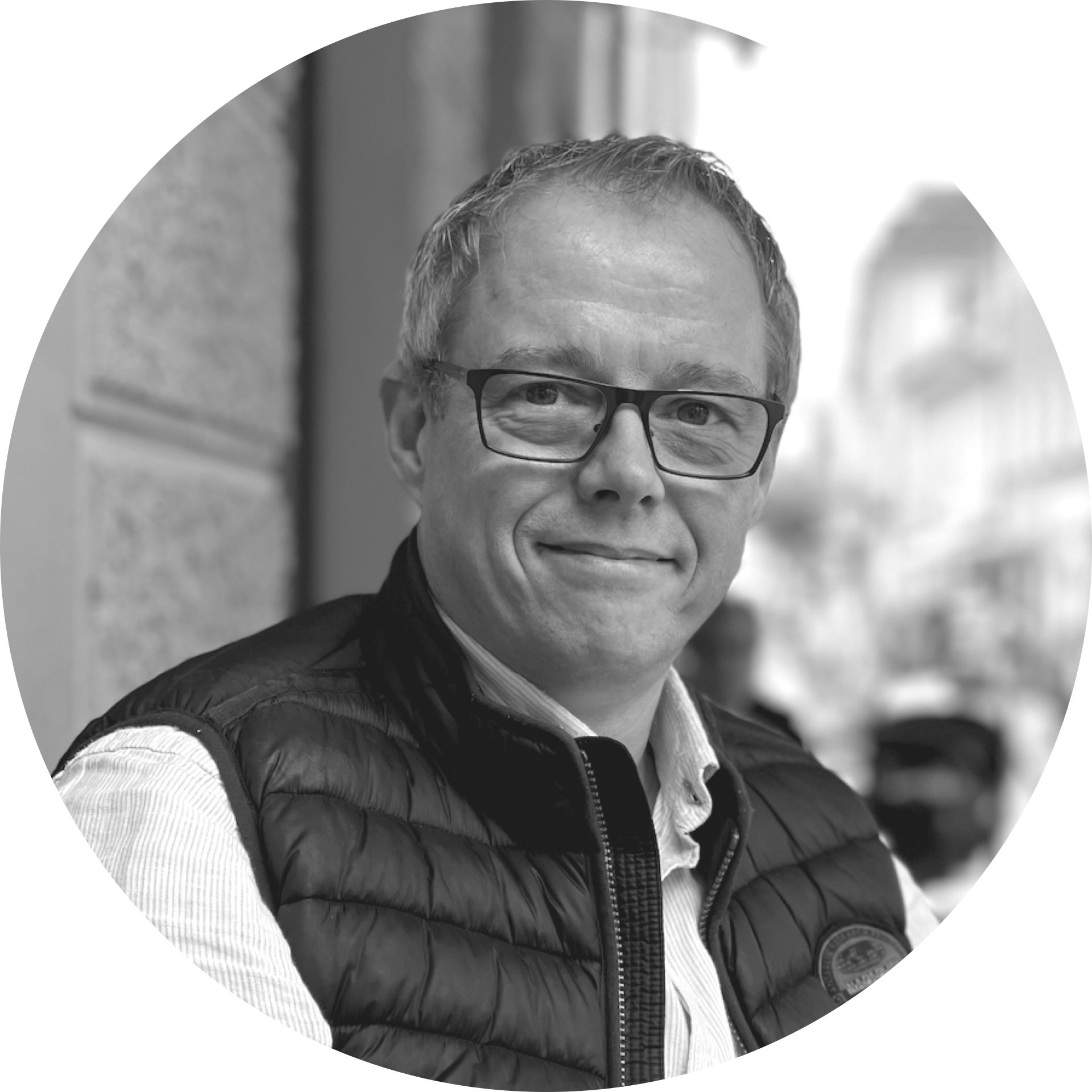 Schwarz-weiß Porträt eines lächelnden Mannes mit Brille, getragen eine moderne schwarze Weste, im Hintergrund eine Stadt mit Gebäuden.