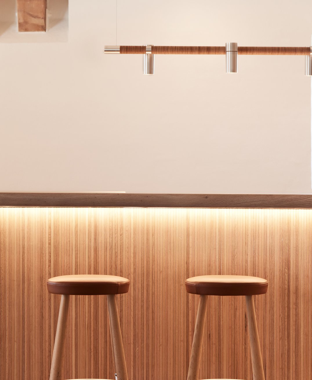 Zwei Holzhocker vor einer Bar mit Holzverkleidung, halbdunkle Wand und minimalistischer Beleuchtung.