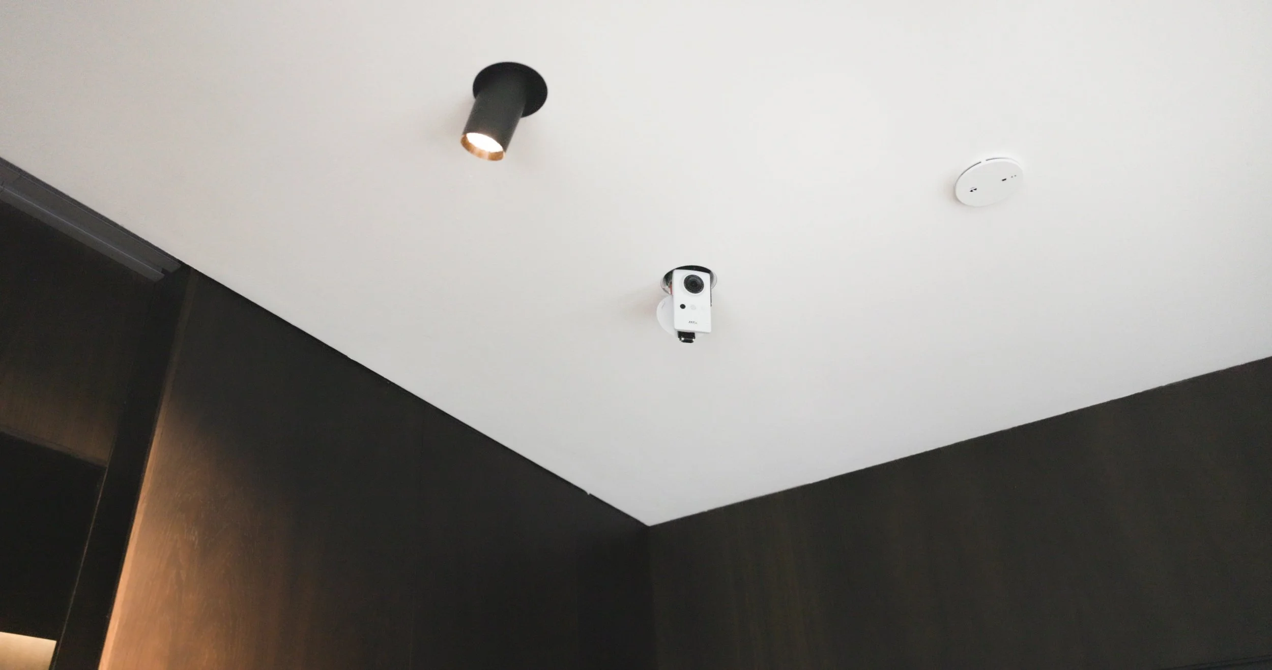 Weißer Überwachungskamera an der Decke, neben einem schwarzen Wandstrahler und einem Rauchmelder in einem Raum mit dunklen Wänden.