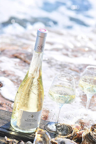 Weißweinflasche an einem Strand mit Muscheln, drei Weinweingläser, Wasser und Meeresküste im Hintergrund